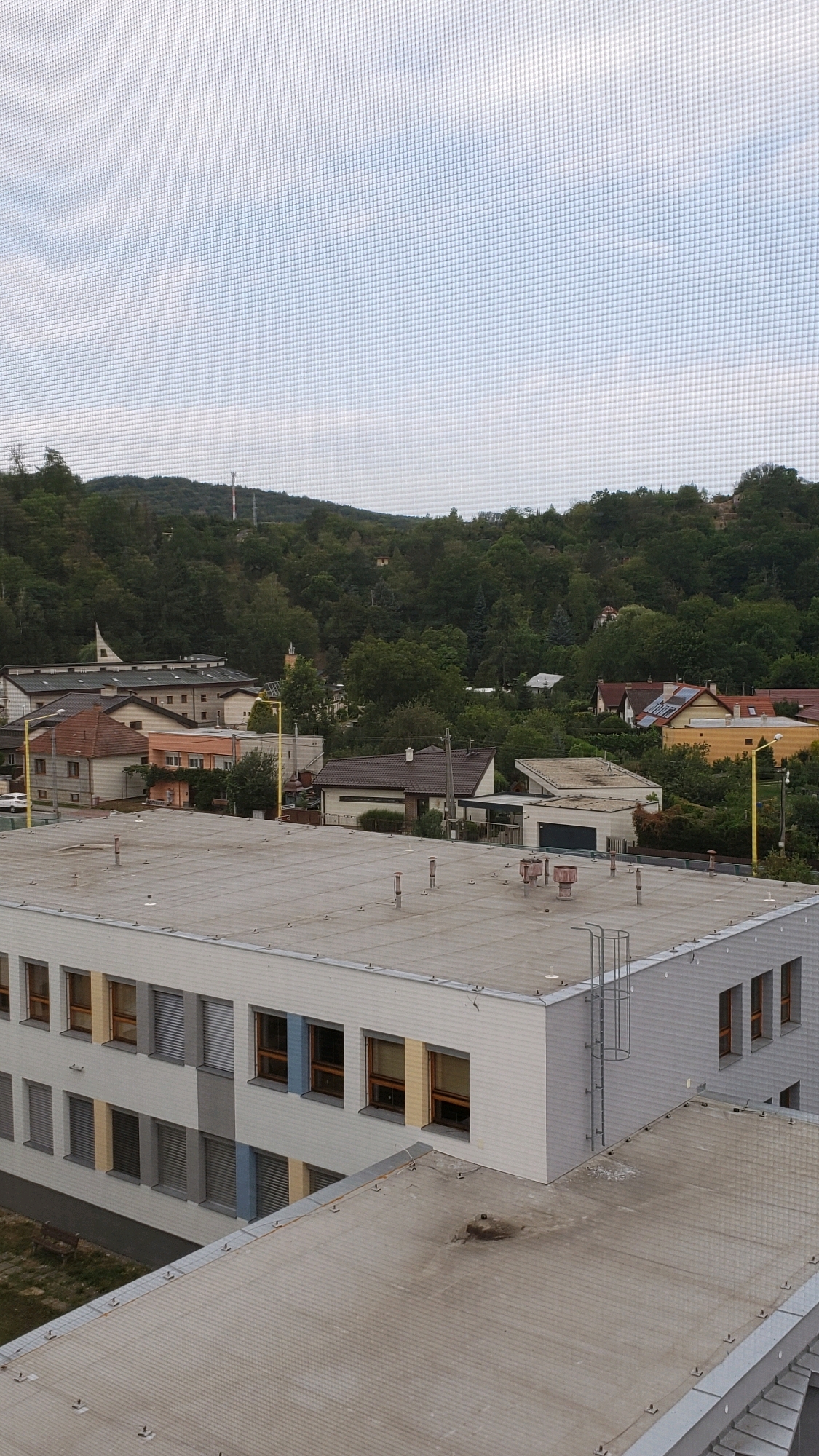 The image is a high-angle, outdoor, daylight shot taken from a building, looking out over a residential area and a wooded hillside. In the foreground is the flat, light-colored roof of a modern, multi-story building, likely a school or public institution, with visible vents and fixtures. Adjacent to this building is another roof, also flat and gray, with a few scattered items and a metal ladder extending up the side of the foreground building.
Beyond the immediate buildings, a collection of houses with various roof types and colors are visible, nestled among mature trees. Some houses have solar panels. Further in the background, a densely wooded hill rises, with a telecommunications tower visible on its summit. The sky is overcast with a grid pattern superimposed on the image, suggesting it was taken through a window screen.
The scene is consistent with the landscape found in the city of Prešov, Slovakia, particularly in areas with a mix of urban development and natural surroundings. The architectural style of the foreground building is characteristic of public buildings constructed in recent decades. The general appearance suggests a typical suburban or semi-rural setting within the city. There are no people visible in the image.