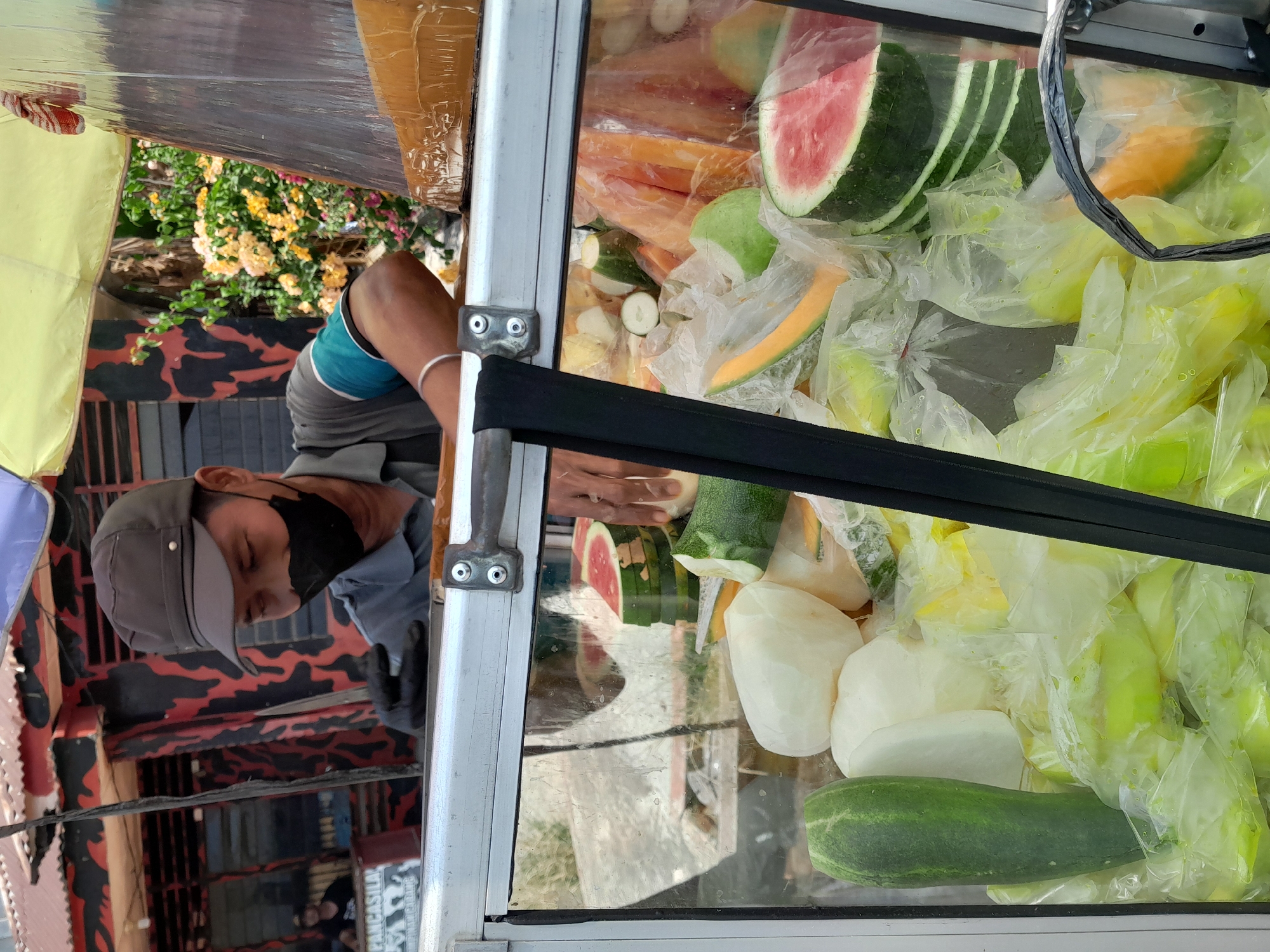 In the city of Kotapinang, Indonesia, a vendor wearing a black face mask and a dark cap is preparing fruit. He is standing behind a glass-fronted cart filled with various fruits. The cart displays several slices of watermelon, some whole cucumbers, peeled white radishes or jicama, and pieces of melon, all packed in clear plastic bags. The vendor is holding a knife and appears to be in the process of cutting or arranging the fruit. In the background, there is a building with a red and black patterned facade and a corrugated metal roof, suggesting a roadside stall or market setting. The scene is illuminated by natural daylight, with a yellow umbrella providing shade over part of the vendor's stall.