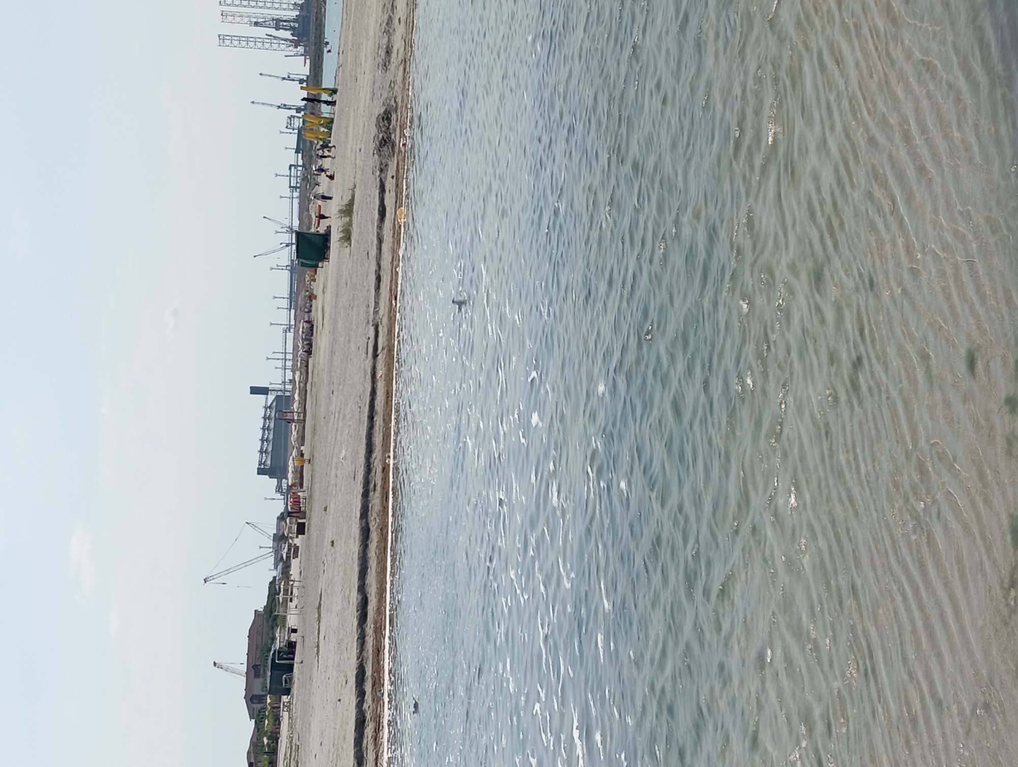 This image depicts a beachfront scene in Ömirzaq, Kazakhstan. In the foreground, shallow, calm water with ripples and some foam covers the sandy bottom. The water's edge meets a light-colored sandy beach, where several small green structures, possibly changing rooms or storage units, are visible. Further back on the beach, a group of people are scattered, some walking and others appearing to be relaxing. Beyond the beach, a distant industrial landscape is evident, characterized by numerous tall cranes, some with long booms, and large industrial buildings, suggesting a port or construction area. The sky is bright and clear, with a few faint clouds. The overall impression is of a functional coastal area that also serves as a recreational space.