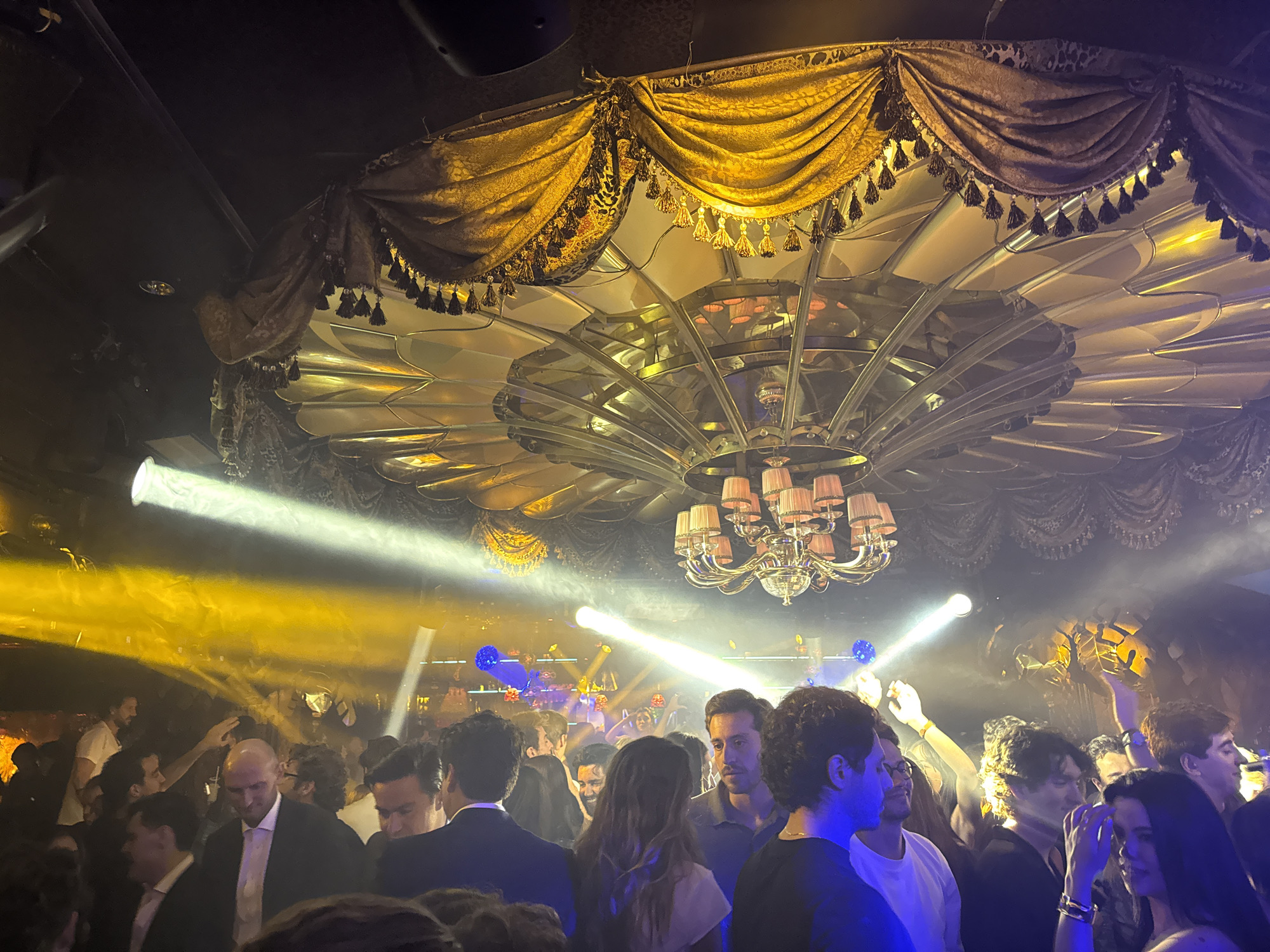 A photograph taken indoors shows a crowded room in Mexico City, Mexico, that appears to be a nightclub. The scene is dimly lit with multiple spotlights and a chandelier hanging from a ceiling adorned with ornate, patterned gold fabric with tasseled edges. The lights create a hazy atmosphere filled with smoke or fog.
Numerous people are present, predominantly facing toward the center of the room. The individuals seem to be dressed in evening attire, with some in suits or dresses. Their activities include talking, standing, and raising their hands in the air. The interactions between the people suggest a social and lively environment.
