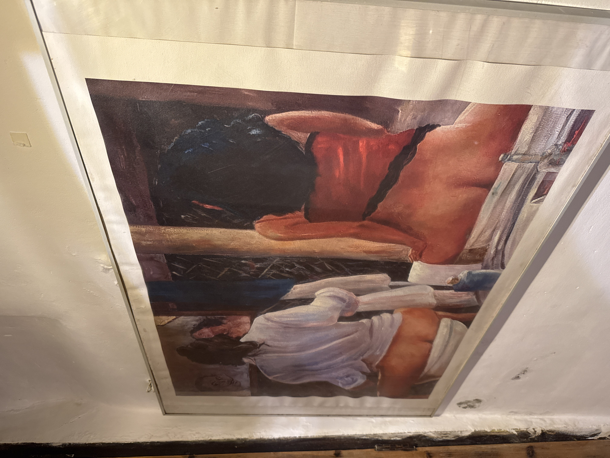 The image shows a framed painting hanging on a white wall, likely in a building in Deià, Spain. The painting depicts a scene of two people, one seemingly lying on a surface with dark hair and the other standing, perhaps leaning over them. The subjects are rendered in shades of brown, red, and blue, and appear to be partially nude. There is the suggestion of a window with curtains to the right of the standing figure. The room's environment appears shadowy and ambiguous. The painting is in a silver frame, and the wall surrounding it shows signs of age and wear. The framing and hanging position suggest the artwork is part of an exhibition or private collection.