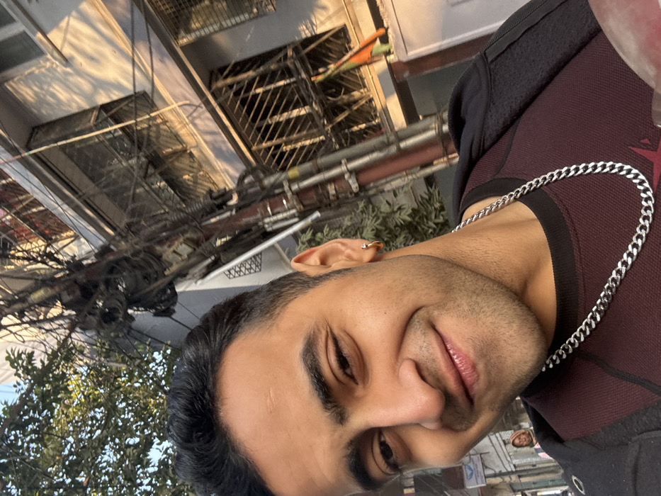 A man smiles into the camera, wearing a maroon top with black accents and a silver chain. An earring is visible in his left ear. Behind him are buildings with caged windows, a tangle of wires, and green trees. The picture appears to be taken outdoors, with a slight tilt.