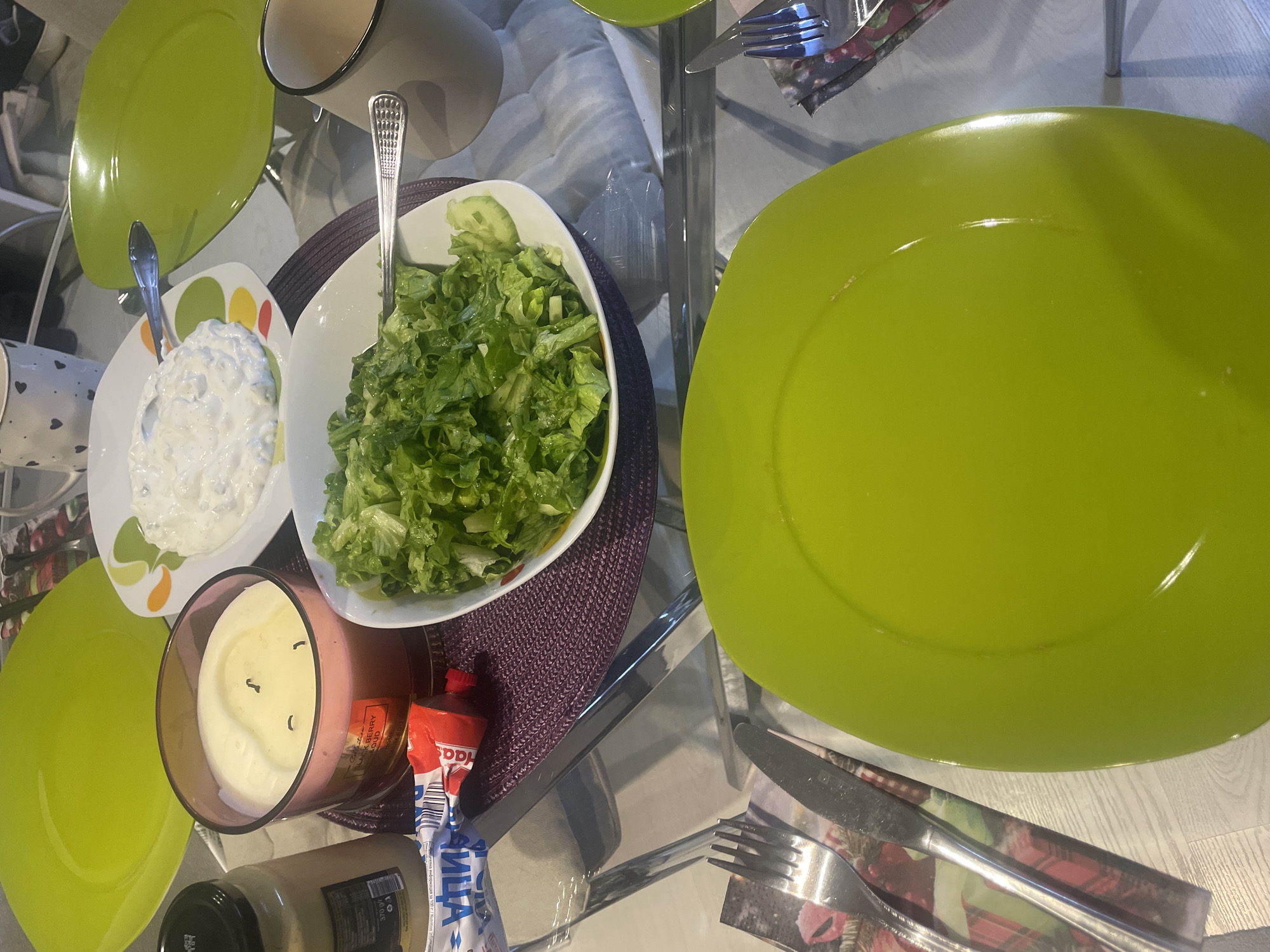 The scene depicts a domestic dining table setting in Sofia, Bulgaria, prepared for a meal. The primary surface is a clear glass table, reflecting overhead light and surrounding elements.
No individuals are present or visible.
The table holds several objects and food items. In the foreground, a prominent lime-green, oval-shaped plate rests on a white surface, with a silver fork and knife placed to its left on a festive-themed paper napkin (red and green design). Another identical lime-green plate is visible further back on the left. A third similar plate is partially visible on the right.
Central to the image, a large white bowl contains a serving of fresh green leaf salad, including visible cucumber slices, with a silver fork resting in it. Adjacent to the salad bowl, on a round, purple woven placemat, is a large, three-wick candle in a pink-tinted glass jar, labeled "Blackberry Cloud." The candle wicks are unlit. To the left of the candle, a beige jar with a black lid displays a label with "370 gr" and Cyrillic text, containing an unidentifiable opaque substance. A partially squeezed red and white tube, bearing Cyrillic text including "МАЙОНЕЗА" (Mayonnaise), rests next to the candle on the placemat.
Further back, a white, rectangular plate with a colorful rim (featuring abstract green, yellow, and red shapes) holds a white, creamy substance that appears to be a dip or spread, with a silver spoon immersed in it. A white mug with black heart patterns is visible in the upper background. To the right, a plain beige mug with a black rim is also present. The overall environment appears to be an indoor setting with ambient lighting.
