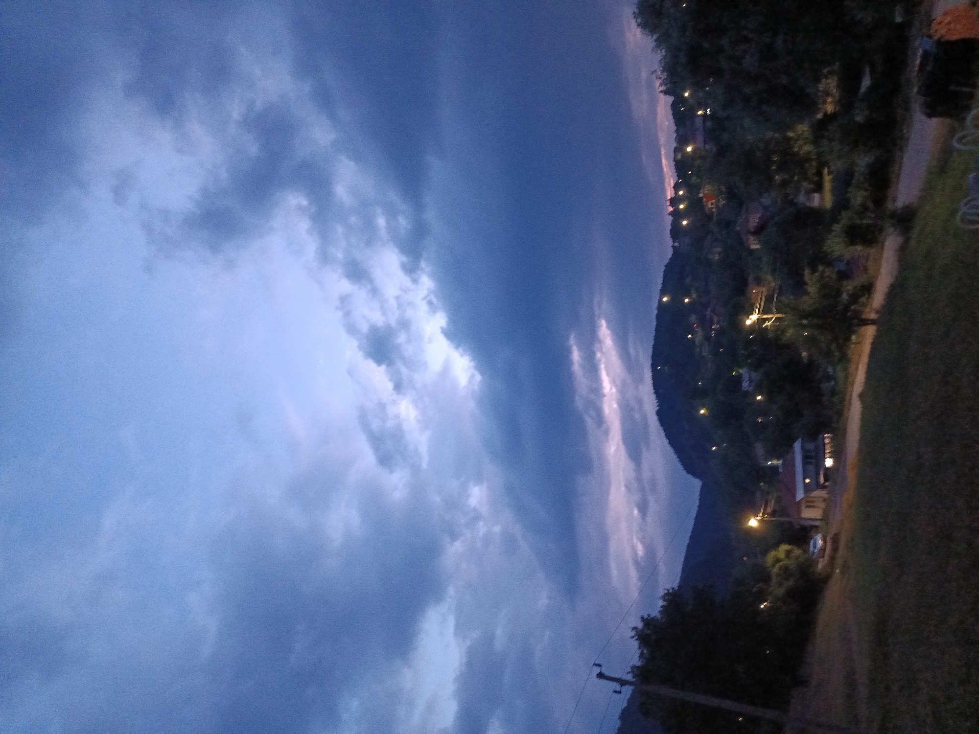 The image depicts a rural village scene in Kralev Dol, Bulgaria, at dusk. The sky is filled with dramatic, dark, and light blue clouds, suggesting an impending or recently passed storm. In the foreground, a grassy slope leads down to a dirt road. A small, light-colored car is parked on the road. To the right of the car is a house with lights on. Beyond the road, a village is nestled on a hillside dotted with more houses, each with illuminated windows. Streetlights are visible throughout the village, casting pools of light. Dark trees and hills form the backdrop, with the overall impression being one of a quiet, residential evening in a mountainous region. No people are visible in the image.