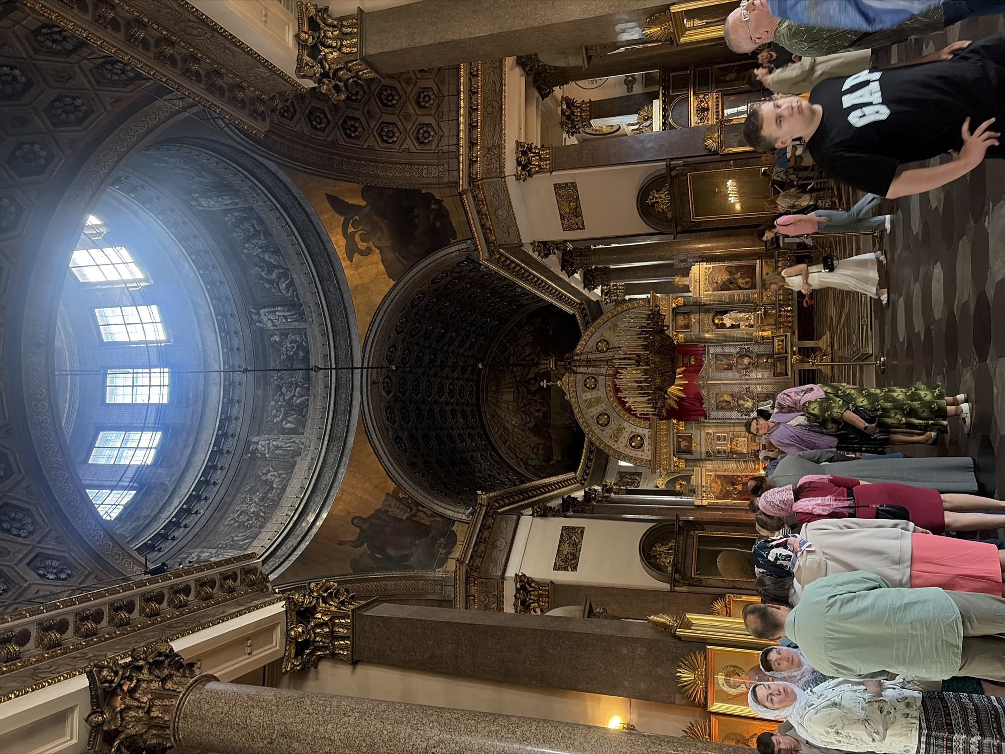 An interior view of a large, ornate cathedral or church features a group of visitors standing and walking within the space. The architecture is grand, with a high, domed ceiling adorned with intricate paintings and geometric patterns. Large, Corinthian columns with gold detailing support the structure. Natural light streams in from windows in the dome, illuminating the interior. Several people are visible, some wearing headscarves, suggesting religious observance. Icons and religious artwork are displayed on the walls. In the foreground, a young man in a black t-shirt with the "GAP" logo stands facing away from the camera. The floor is made of patterned stone tiles. The overall impression is one of solemnity and historical grandeur.