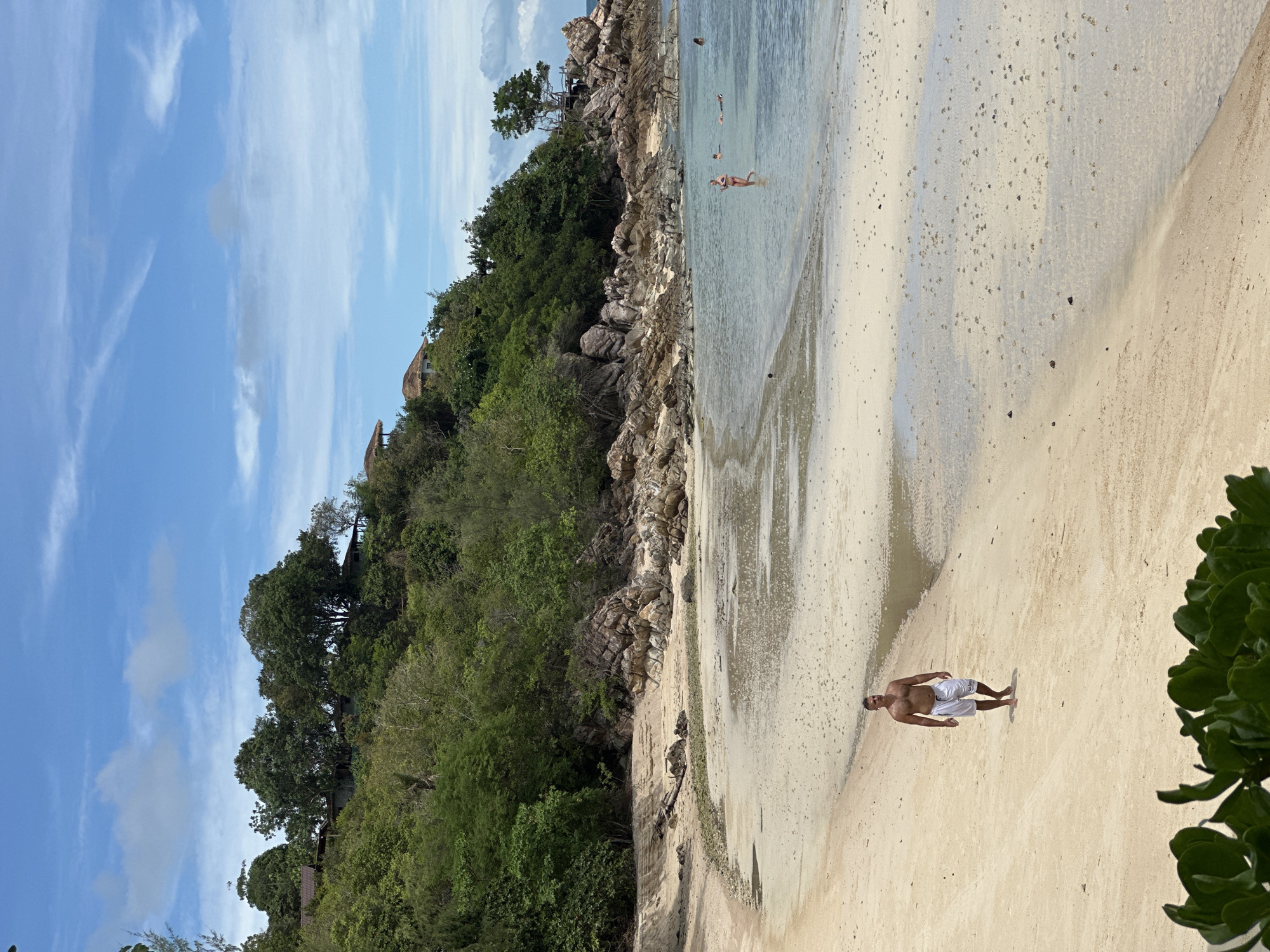 A man in white shorts walks across a sandy beach on Koh Phangan, Thailand. He is walking away from the water and towards the viewer. The beach is wide and the sand is light brown. In the background, there are rocks and lush green trees on a hillside. The water is clear and shallow, with a few people swimming. The sky is blue with some white clouds.