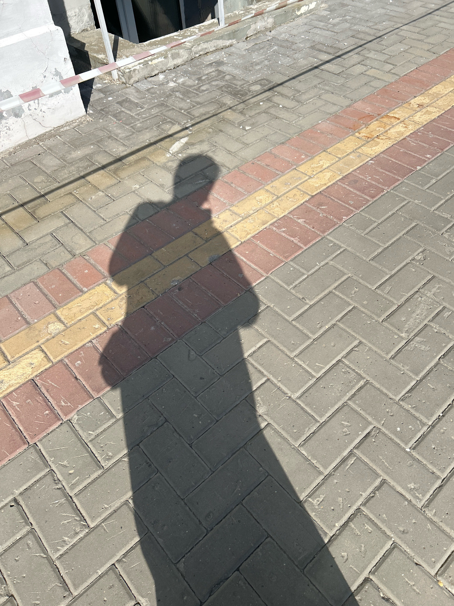 An outdoor shot shows a shadow of a person on a paved walkway. The walkway is made of interlocking bricks arranged in a herringbone pattern. There are strips of red and yellow bricks interspersed with the grey bricks, creating a visual border. The lighting suggests it is daytime, casting a long shadow diagonally across the scene. A white wall with some debris is visible in the upper left corner, along with a red and white striped tape barrier. The environment appears to be an urban or pedestrian area. The image does not contain any specific details to indicate it is located in Tomsk, Russia.