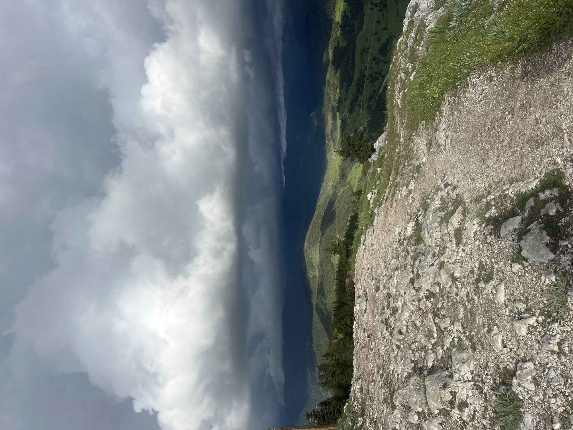 The image presents a landscape view from an elevated perspective, likely a mountain or high ridge. A rugged, rocky terrain dominates the foreground, transitioning into a grassy edge. In the middle ground, rolling hills covered with green vegetation and dark patches of forest stretch into the distance. A prominent dark, ominous storm cloud formation hangs over the landscape, casting a shadow on parts of the hills, while brighter, white clouds are visible in other sections of the sky. The location is speculated to be in or near Iagodina, Bulgaria, based on provided information. No people, actions, or specific interactions are discernible in the image.