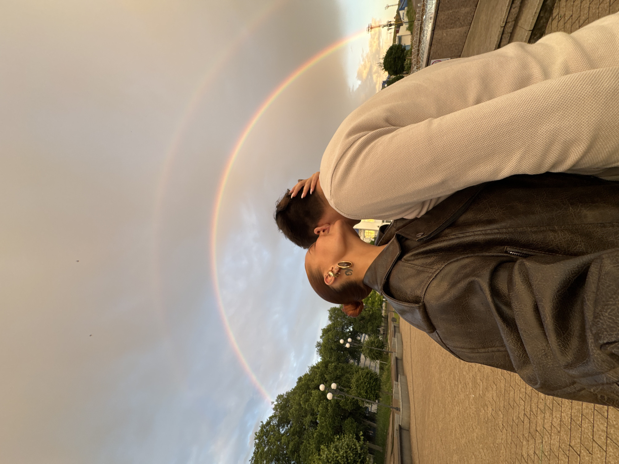 A couple kisses in front of a double rainbow in Chaikovsky, Russia. The woman, on the left, is wearing a brown leather jacket and large hoop earrings. Her hair is pulled back in a bun. The man, on the right, is wearing a light-colored sweater. They are standing on a paved area with trees and lampposts in the background. The sky is cloudy with a prominent double rainbow.