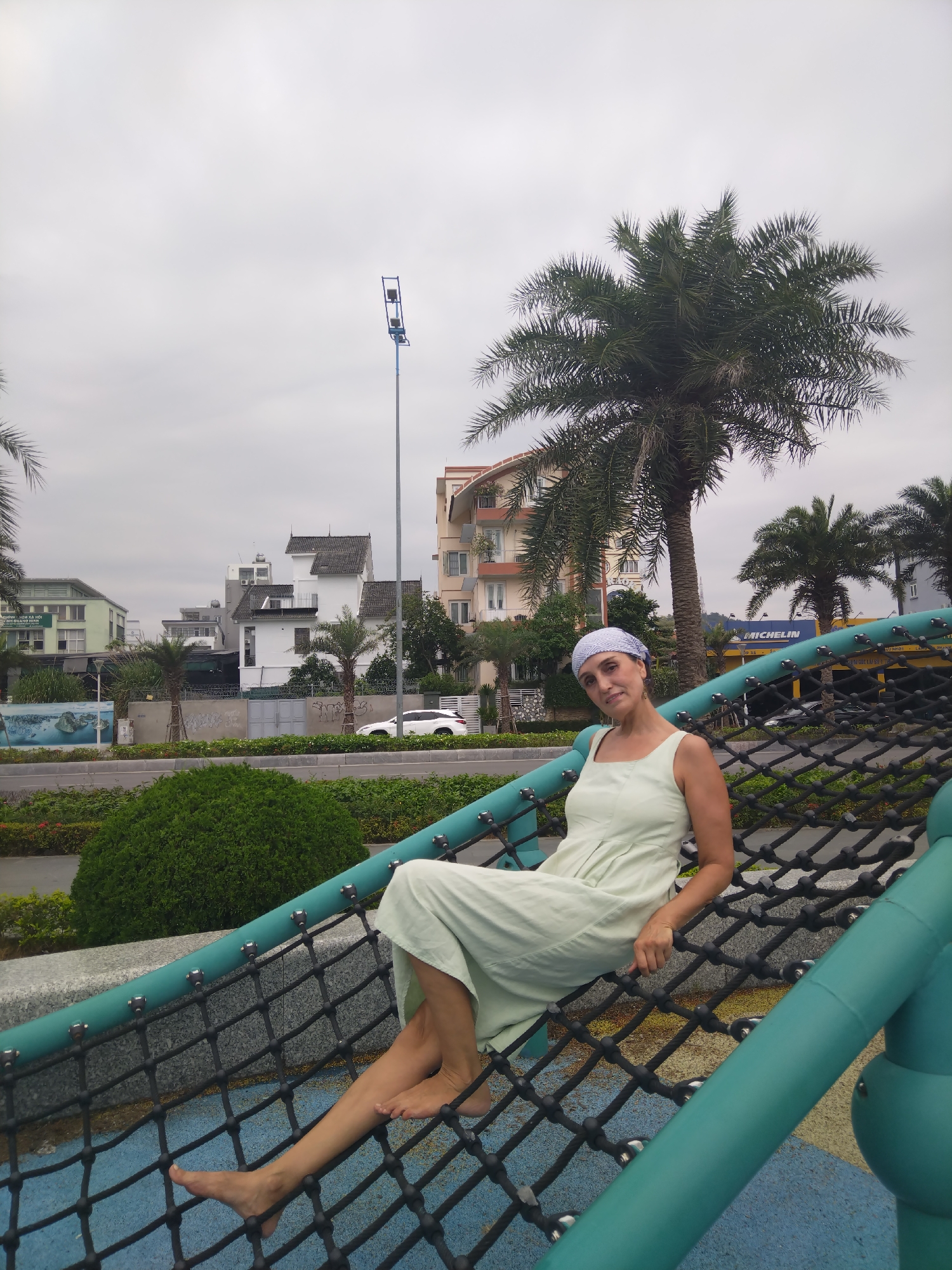 A woman wearing a light green dress and a patterned headscarf sits on a black rope net structure with green tubular supports. She is barefoot and leans slightly back, looking towards the camera. The background includes palm trees, buildings, and street vegetation. The location appears to be in Ha Long Ward, Vietnam, based on architectural styles and signage. A tall blue streetlight is visible in the distance.
