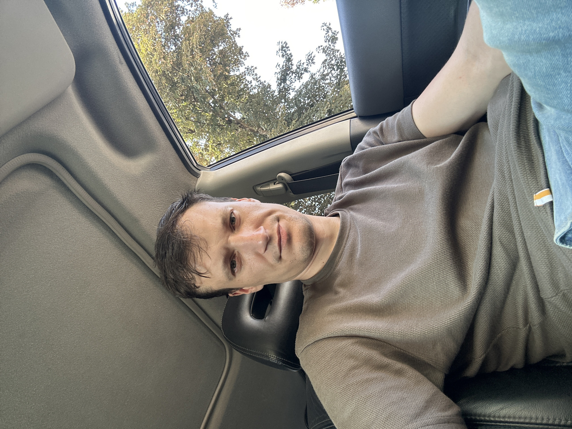 A man is sitting in the passenger seat of a car, looking directly at the camera. He is wearing a brown, long-sleeved shirt and blue jeans. The interior of the car is visible, including the black leather seat and headrest, and the grey headliner. Through the window, a tree with green and yellow leaves can be seen, suggesting an outdoor setting. The image appears to have been taken during the daytime, with natural light illuminating the scene. There is no specific indication of the location within Kamianets-Podilskyi, Ukraine.