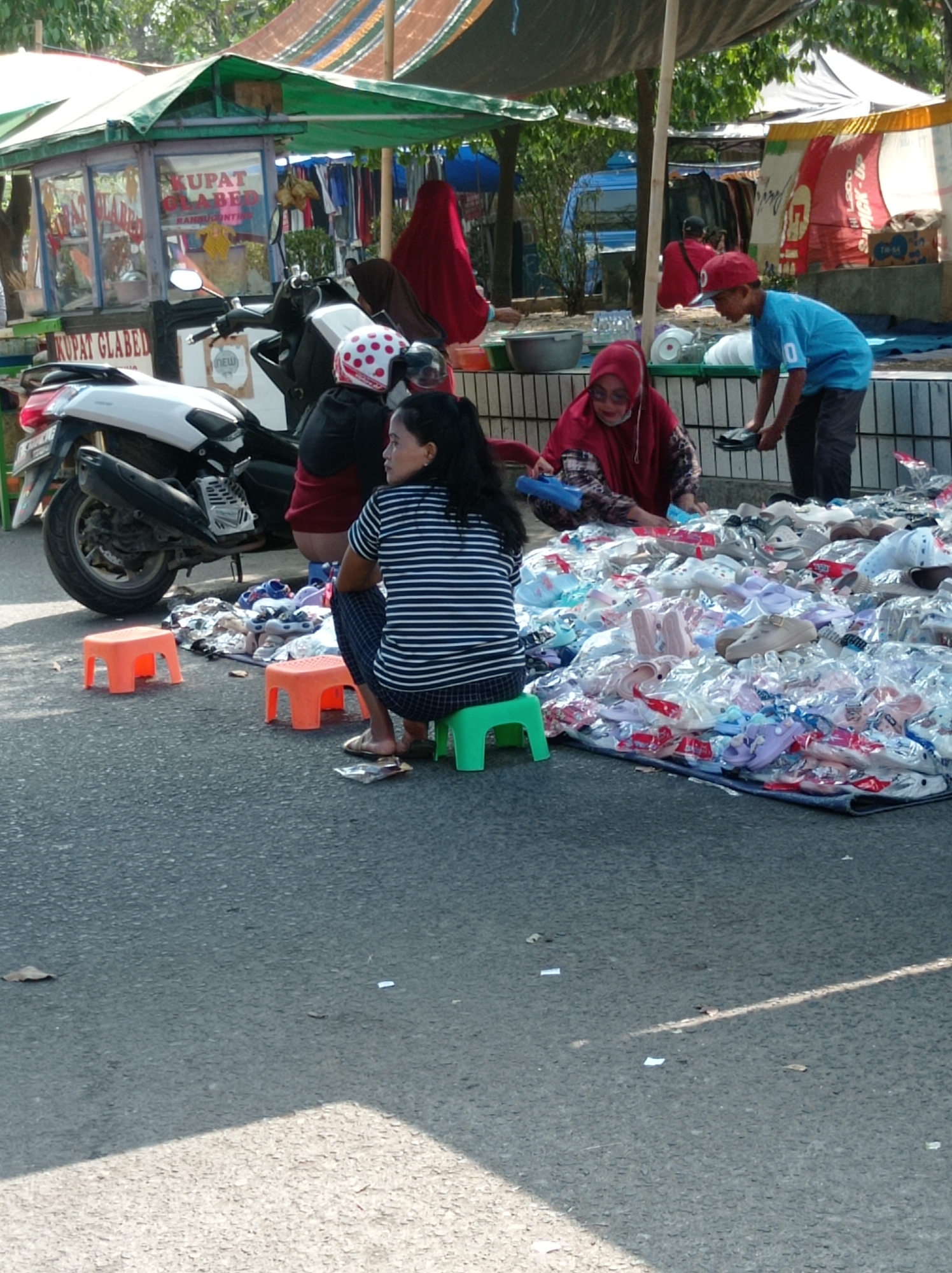 The image depicts a street-level view of a market stall in Tegal, Indonesia, likely selling footwear. In the foreground, a woman wearing a striped t-shirt and dark pants is seated on a green stool, facing away from the camera. Next to her, on the right, a large tarp is spread on the ground, covered with a substantial quantity of footwear, including sandals and shoes, many of which appear to be packaged.
To the left of the woman, two small orange stools are placed on the asphalt. Behind these stools and the woman, a white motorcycle is parked. Further back, a food stall with a green awning displays signage that reads "KUPAT GLABED." A woman wearing a red headscarf and patterned clothing is crouched behind a white plastic tub, interacting with a young boy. The boy, wearing a blue t-shirt and a red cap, is also crouched and appears to be handling items from the tarp.
In the background, a red awning provides shade over a portion of the market area. Other stalls and market activity are visible, with various fabrics and goods displayed. The overall scene suggests a bustling outdoor market environment.