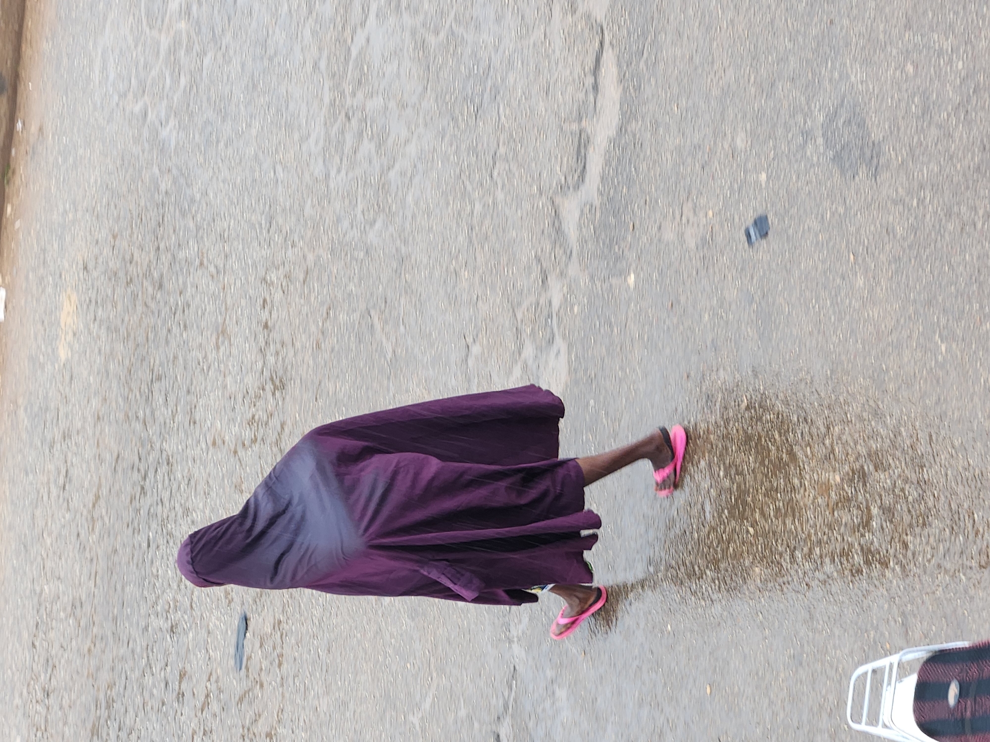 A person wearing a purple head covering and a dark purple skirt walks on a wet, paved surface. The person wears pink flip-flops. The surface appears to be a road or sidewalk with puddles reflecting the overcast sky. A part of a white and black object, possibly a scooter or bicycle carrier, is visible in the lower left corner. The background is out of focus and shows the texture of the paved surface.