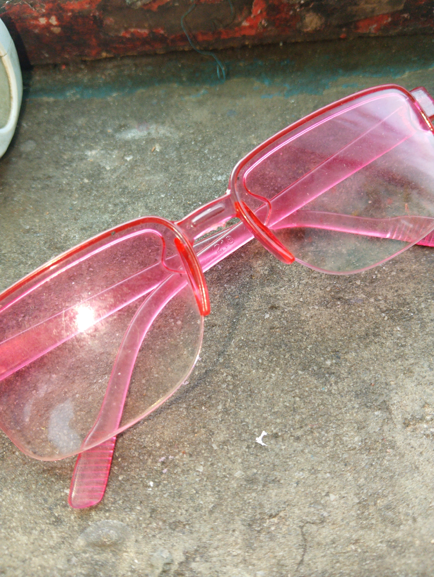 A pair of pink plastic framed glasses lies on a rough concrete surface. The glasses are angled, with the lenses facing upwards and slightly to the right. The frames are a translucent pink, and the lenses appear to be clear, though they are covered in dust and smudges. The concrete surface shows signs of wear and tear, with cracks and small stones embedded in it. In the background, a weathered red and black surface is visible, possibly part of a wall or barrier. A small white object, possibly a piece of paper, is also present on the concrete near the glasses. The overall scene suggests an outdoor environment, potentially in an urban setting like Lagos, Nigeria, where such discarded items are common on public walkways or neglected areas.