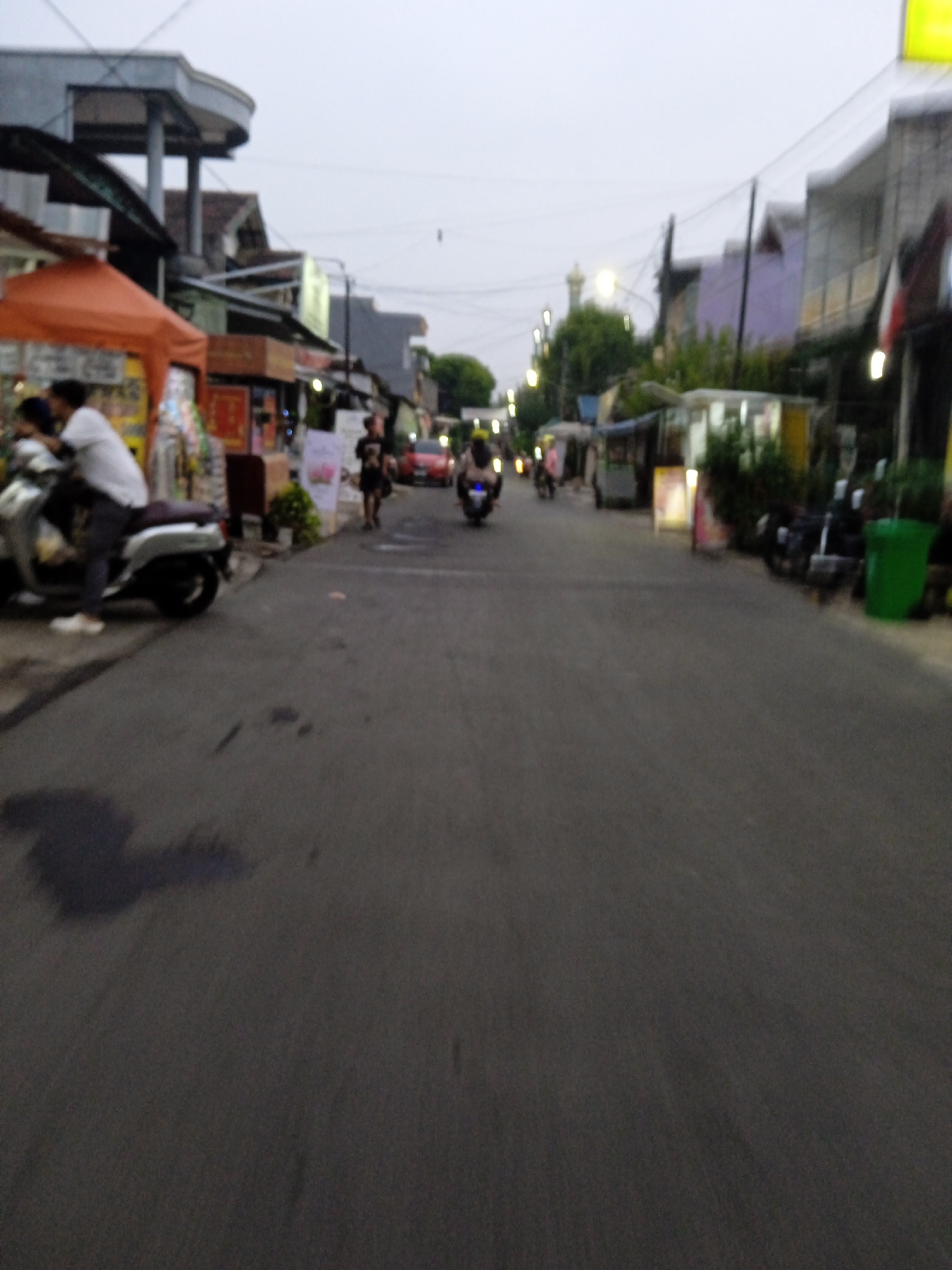The image depicts a street scene in Bekasi, Indonesia, likely during the late afternoon or early evening, as indicated by the diffused lighting and artificial streetlights. The perspective is from a low angle, suggesting the photo was taken from a vehicle or while walking.
The foreground is dominated by a gray asphalt road with some water stains or dark patches. The middle ground shows a bustling street lined with shops and food stalls. On the left, an orange awning shelters a food stall with people gathered around it. A white motorcycle is parked to the left, with two individuals on it, one wearing a white shirt and the other a dark shirt. Further down the street, a few more motorcycles are visible, some with riders, suggesting ongoing traffic and activity.
On the right side of the street, there are buildings with various facades, some appearing residential or commercial. One building has a distinctive light purple wall. A bright green trash bin is prominently placed near the right edge of the frame. Streetlights are visible along the road, casting a warm glow. In the distance, a mosque minaret can be seen, a common architectural feature in Indonesian cities.
The overall impression is of a typical urban environment in Bekasi, characterized by everyday commerce, transportation, and community life. The image captures a moment of ordinary activity, with people engaged in shopping, commuting, and socializing.
