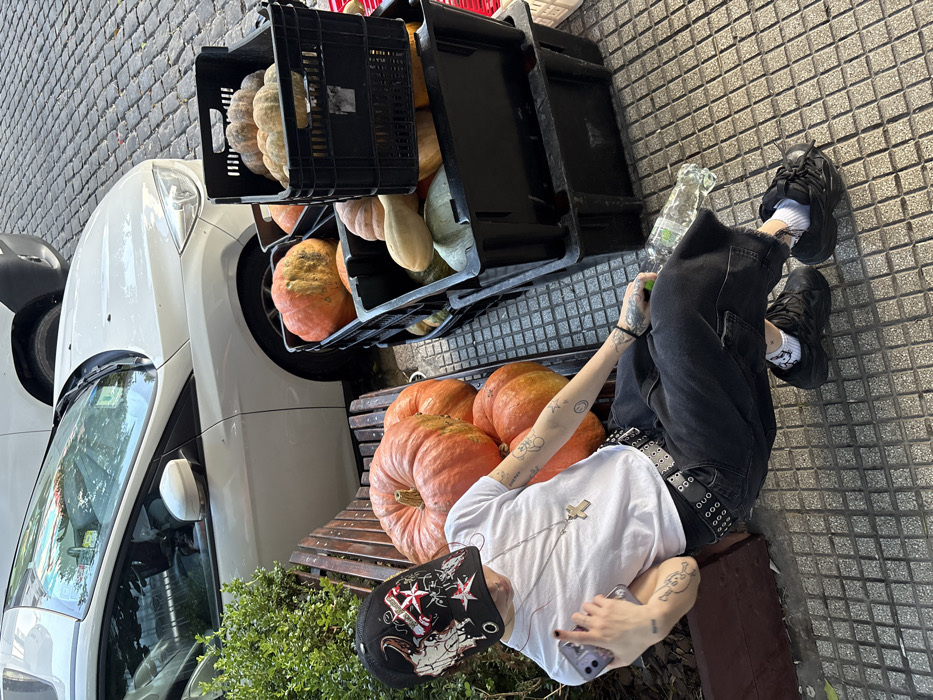 A person sits on a wooden bench beside a stack of black crates filled with various pumpkins. Several large orange pumpkins rest on the bench around the person. The individual wears a white t-shirt, dark jeans with a studded belt, and black sneakers with white socks. A black patterned cap sits on their head. A car is parked to the left of the bench, and tiled pavement is visible on the right.