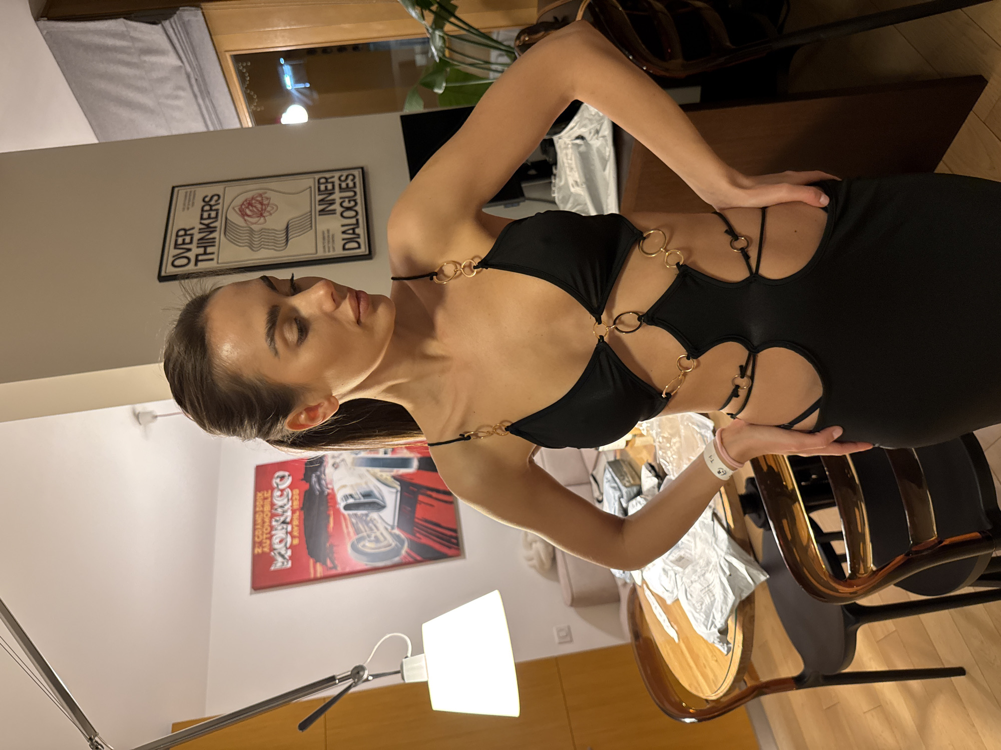 A woman with long brown hair, wearing a black dress with ring detailing, is posing indoors. She has a white bracelet on her left wrist. The location seems to be an apartment, evidenced by the presence of furniture, posters, and a lamp. Two posters are visible: one with the text "OVER THINKERS INNER DIALOGUES" and another with racing-themed imagery with the word "MONACO." The location is most likely Warsaw, Poland, but there's no explicit evidence visible to verify this claim.