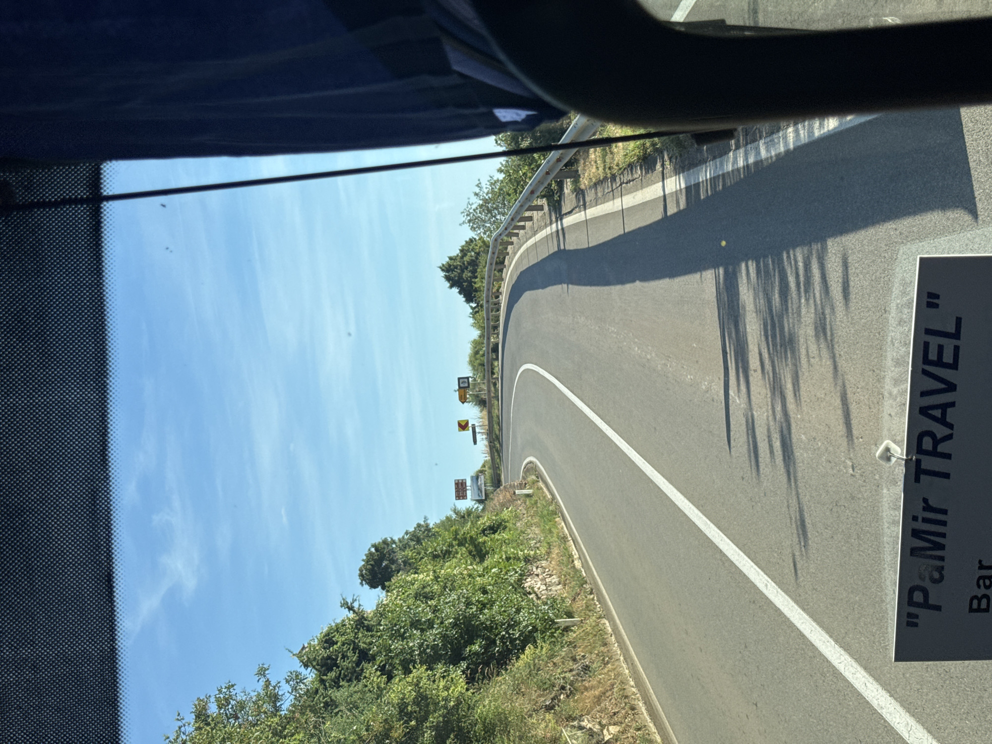 The image captures a view from a vehicle, likely a bus, traveling on a winding road in the city of Blizikuće, Montenegro. The road curves to the left, bordered by a metal guardrail on the right and a steep, tree-covered embankment on the left. The sky above is a clear blue with a few wispy clouds. Road signs are visible in the distance, indicating curves in the road. The lower portion of the image shows a sign that reads "\"PaMir TRAVEL\" Bar". The overall scene depicts a scenic mountain road in a coastal region, suggesting a tourist or travel context.