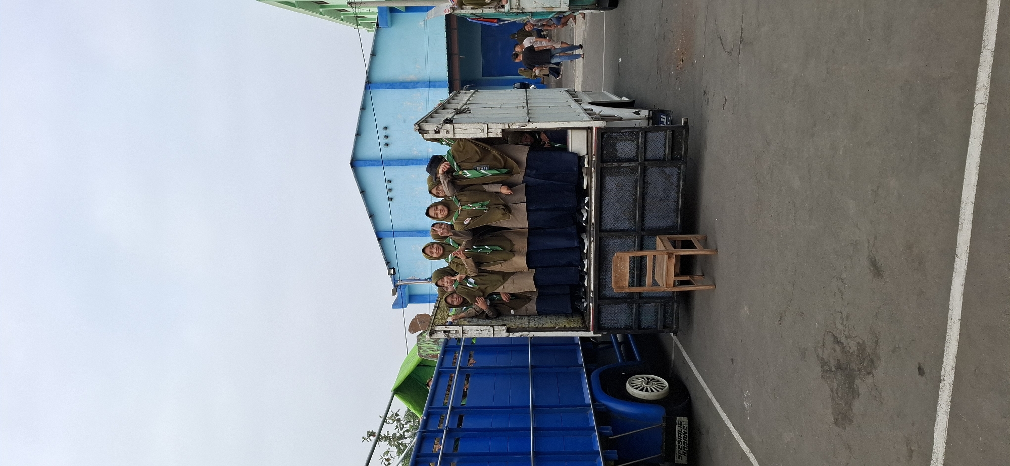 In Wonorejo, Indonesia, a group of young women in matching brown scout uniforms and green scarves are gathered in the back of a white truck. The truck is parked next to a larger blue truck, and the women are standing closely together, many of them smiling and some posing with hand gestures. A simple wooden chair sits on the floor of the white truck in front of the group. In the background, a blue building with a corrugated roof is visible, and further beyond, a green building. Several other people are walking in the distance. The scene appears to be outdoors on an overcast day, with the ground being a grey, cracked asphalt surface marked with white lines.