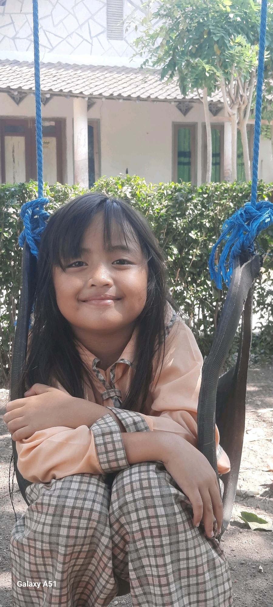 A young girl with dark hair is smiling while sitting on a swing. She is wearing a light peach colored collared shirt with plaid trim on the cuffs and a plaid skirt. The swing has thick black sides and is suspended by blue ropes. Behind her is a neatly trimmed green hedge. In the background, a white building with a tiled roof and some trees are visible. The lighting suggests it is daytime. The text "Galaxy A51" is visible in the bottom left corner.