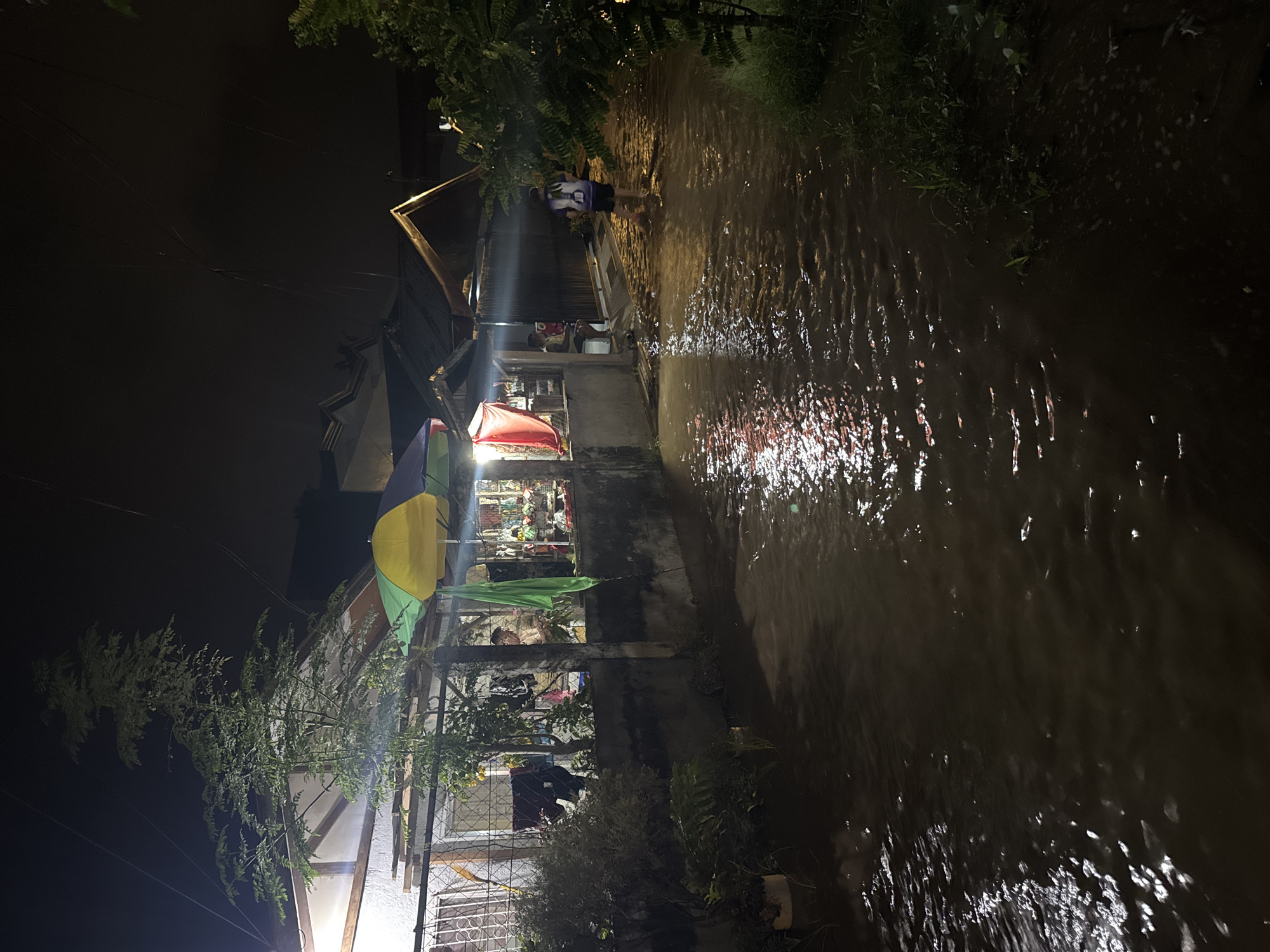 The image captures a flooded street at night in what appears to be an urban or semi-urban area of Davao City, Philippines. The water level is high, covering the road and creating ripples that reflect the lights from nearby structures. On the left side of the frame, a building with a white facade and a sloped roof is visible, with what looks like a small shop or dwelling. Several umbrellas are set up in front of this building, suggesting a market stall or outdoor seating area. A man is seated near the entrance of the building. Further down the street, on the right side, another person is walking through the flooded area, their back to the camera. Lush green vegetation is present on the right side of the street, indicating a tropical environment. The overall scene suggests a community experiencing flooding, possibly due to heavy rainfall, with some economic activity continuing despite the conditions.
