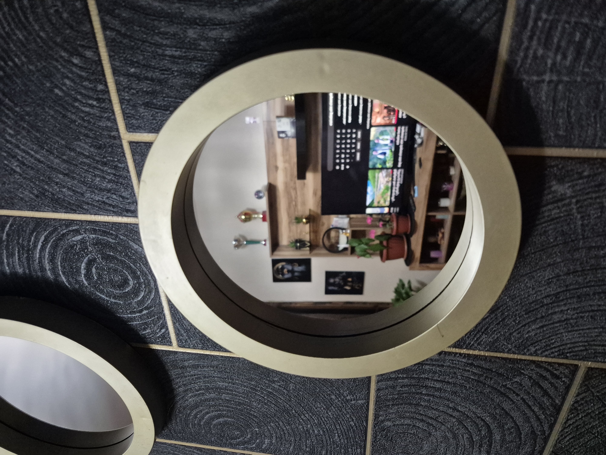 The image shows two round mirrors with gold frames hanging on a dark textured wall with gold-colored grout. The larger mirror reflects the interior of a room containing a television mounted above a wooden shelf unit. The shelf unit contains various decorative items, including plants, trophies, and framed artwork. The scene is likely indoors, potentially in a residence or a shop. The location is Tbilisi, Georgia. There are no people visible, only objects reflected in the mirror.