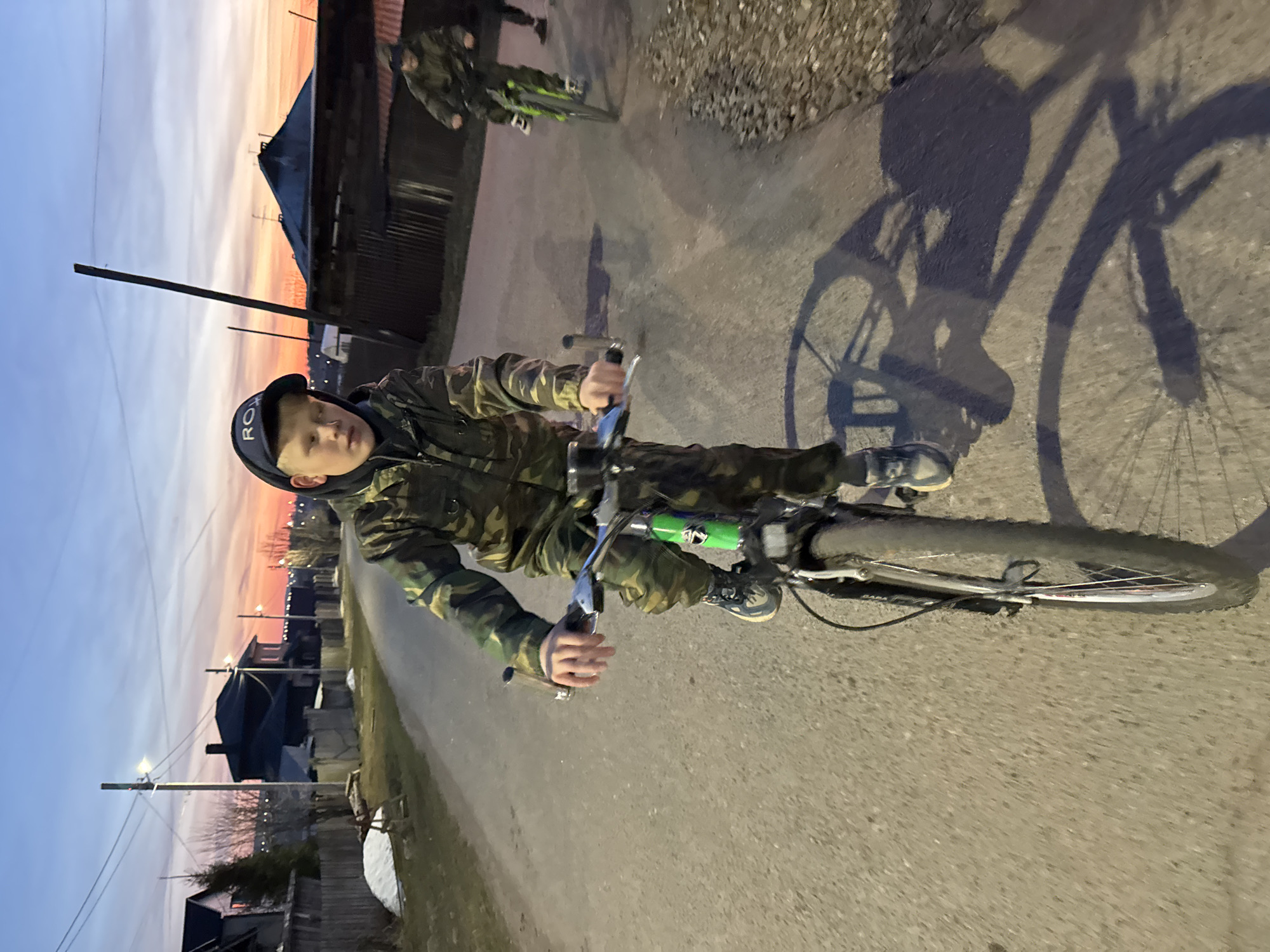 The image is oriented sideways, depicting a rural or semi-urban road scene at dusk. The main subject is a young boy, likely pre-teen, riding a bicycle and looking towards the camera. He wears a camouflage patterned jacket and a dark baseball cap with the white letters "ROY" visible on it.
Further down the road, another person on a bicycle is visible in the background. The road appears to be unpaved or minimally paved. Along the side of the road, there are houses with dark roofs and power poles with illuminated streetlights. Power lines crisscross the upper part of the frame.
The sky, visible in the background, displays warm colors of orange and pink along the horizon, transitioning to light blue higher up, characteristic of a sunset or twilight. Long, distinct shadows of the bicycles and riders are cast on the ground, indicating a low light source. The boy's bicycle features a bright green element on its frame, possibly a water bottle holder or accessory. The setting suggests a small town or village in an area like Горный, Russia.
