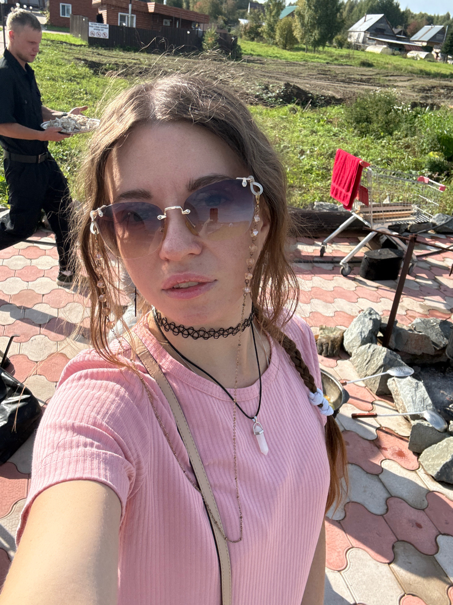 A young woman with long, braided brown hair and wearing a pink ribbed t-shirt and sunglasses is taking a selfie outdoors. She has a choker necklace and a pendant necklace. In the background, a man in a black uniform is holding a plate of food. Behind them, there is a shopping cart with a red towel hanging from it, a fire pit, and some large rocks. The ground is paved with pink and white tiles. In the far background, there are houses and trees, suggesting a rural or semi-rural location in the city of Pakly, Russia.