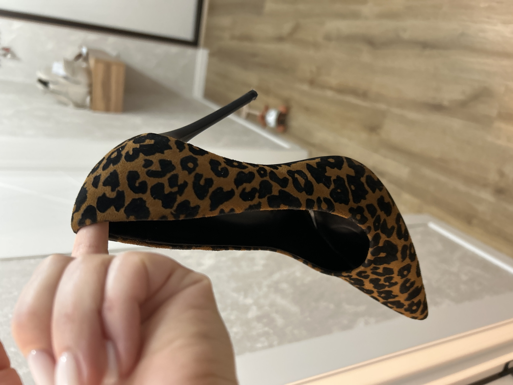 The image displays a close-up, slightly tilted view of a single high-heeled shoe featuring a distinct leopard print pattern. The shoe is a classic pump style with a pointed toe and a thin, black stiletto heel. Its material appears to be suede or a similar textured fabric, patterned with black spots against a tan or light brown background.
A person's hand, with neatly manicured fingernails, is visible in the foreground, holding the shoe. One finger is inserted under the shoe's arch, suggesting it is being held up for examination, to check its flexibility, or to show its design.
The background is softly blurred, indicating an indoor setting, likely a modern home. Visible elements include light-colored countertops or surfaces in the mid-ground, and further back, a wooden wall or flooring with a horizontal grain pattern. Some indistinct, out-of-focus objects, possibly decor or appliances, can be seen on the countertop. The lighting suggests an indoor environment. No text is visible in the image. The scene offers a glimpse into a domestic setting within the context of Chaikovsky, Russia.