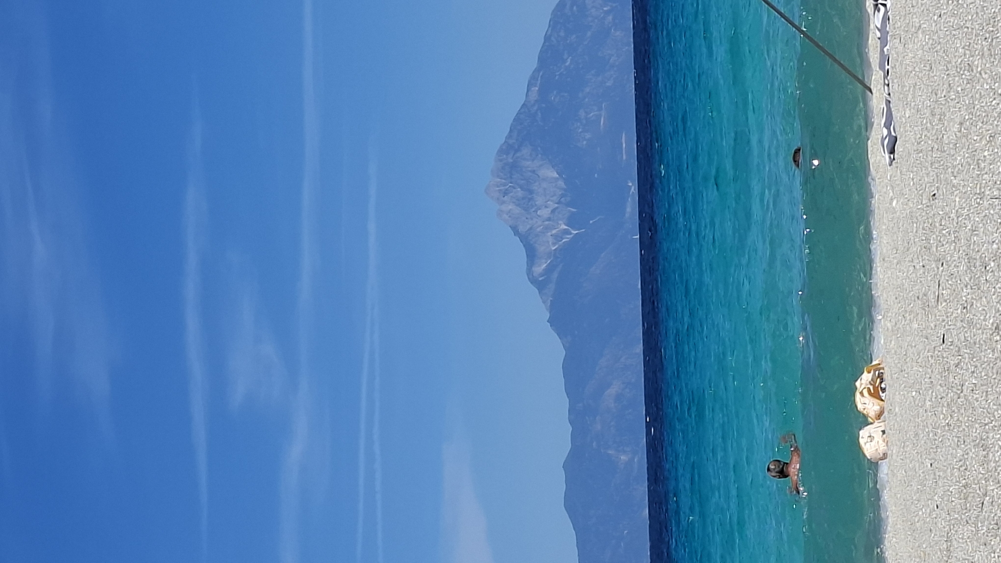 The image depicts a view of the Aegean Sea with a majestic mountain range in the background, likely Mount Olympus. The water is a vibrant turquoise, with a clear blue sky overhead adorned with thin wispy clouds. In the foreground, a sandy beach is visible, with two persons swimming in the sea. One person, partially submerged, appears to be swimming towards the shore, while another person's head is visible in the water. Two rolled-up towels or bags are resting on the beach near the water's edge. The scene suggests a serene day at the beach. The location is a coastal area in the Toroni Municipal Unit, Greece, with the prominent mountain range identifiable as Mount Olympus.