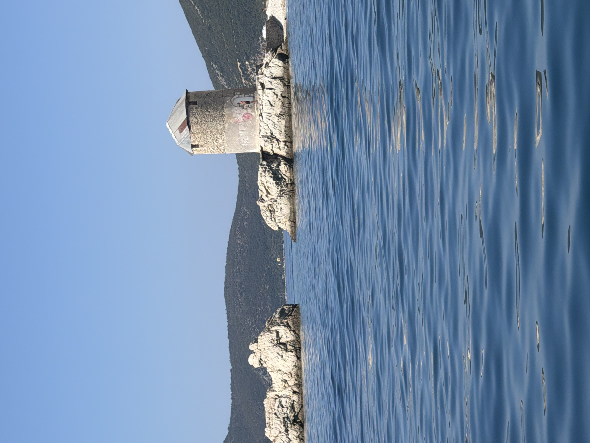 The image captures a picturesque coastal scene in the Municipal Unit of Kalamos, Greece. In the foreground, the deep blue water of the sea is calm, with subtle ripples reflecting the clear blue sky. To the left, two large, light-colored rocky formations rise from the water. In the middle ground, a stone-built tower, likely an old windmill, stands on a rocky outcrop. The tower is cylindrical with a conical, corrugated metal roof. Graffiti is visible on the lower part of the tower. A person, appearing to be a man wearing dark shorts, is standing near the entrance of the tower. In the background, lush green hills covered in vegetation stretch across the horizon, meeting the clear blue sky. The overall atmosphere is serene and idyllic, showcasing the natural beauty of the Greek coastline.