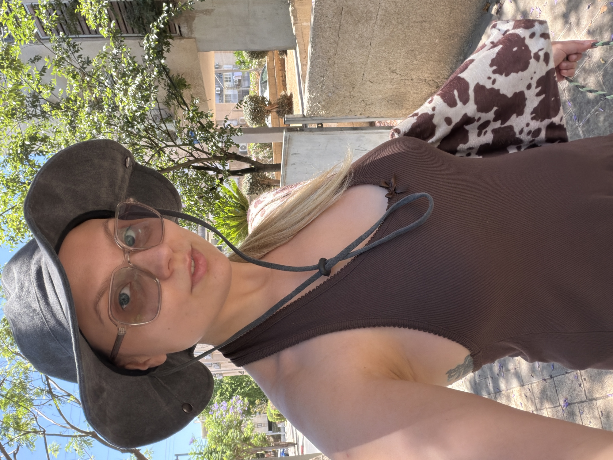 The image, rotated 90 degrees counter-clockwise, presents a close-up outdoor selfie of a young woman. She has light-colored hair and wears rectangular-framed glasses, looking directly at the camera with a neutral expression. She is sporting a dark grey wide-brimmed hat with a chin strap and a brown ribbed tank top or dress. A small tattoo is visible on her left arm. Draped over her right arm/shoulder area is a distinctive white and brown cow-print fabric.
The background suggests an urban environment under bright daylight. Lush green tree foliage occupies the upper part of the scene, with glimpses of clear blue sky. In the mid-ground, residential or commercial buildings are visible, characterized by their light-colored facades and external air conditioning units. Fences or railings and small manicured bushes line the area. The ground beneath is paved with square-patterned stones, upon which scattered purple floral debris is visible. The strong sunlight casts sharp shadows, indicating a sunny day, likely morning or afternoon. This outdoor setting, with its specific architecture and flora, aligns with locations found in cities like Tel-Aviv, Israel. No visible text is present.