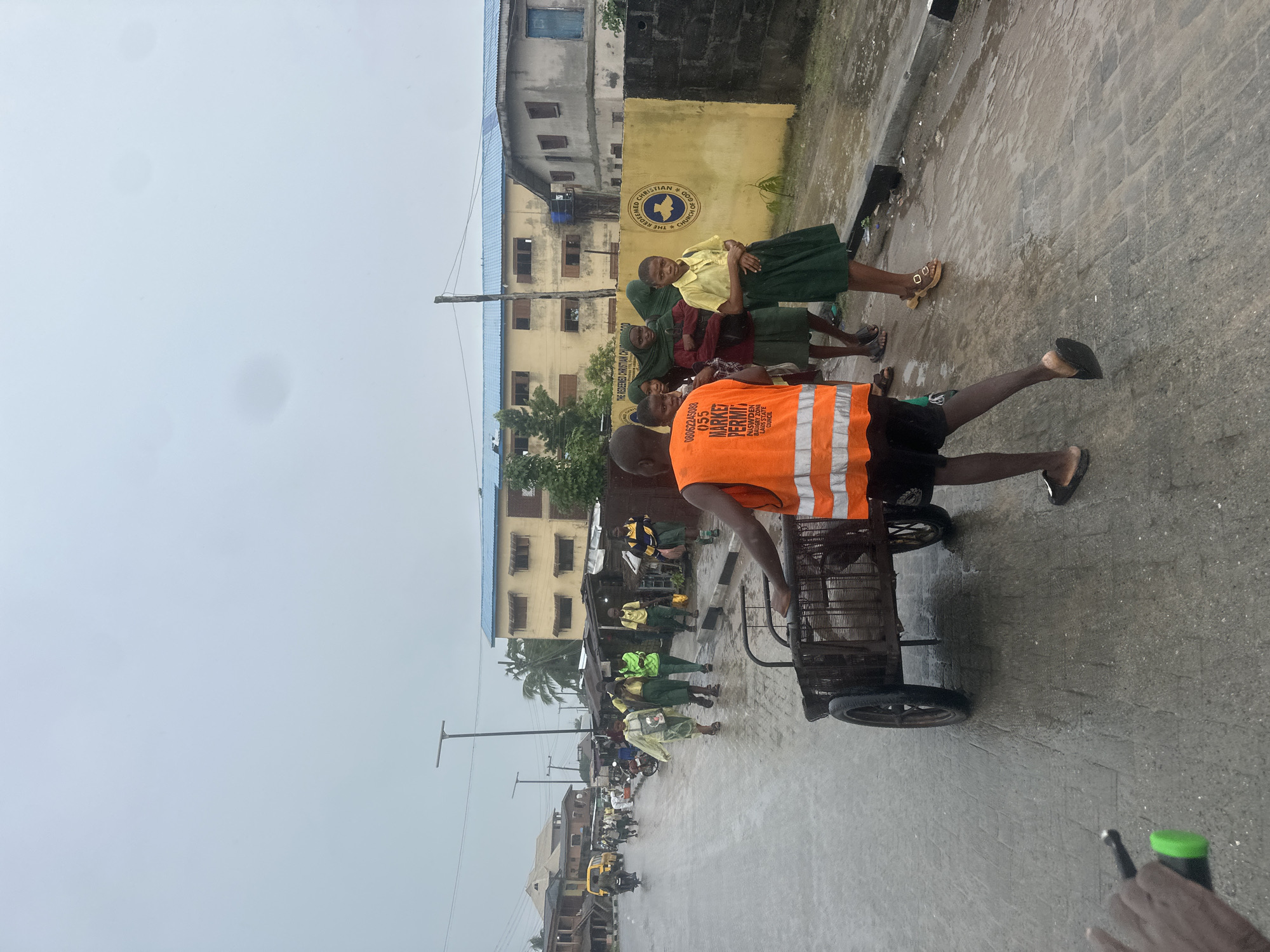 The image depicts a street scene in Badagry, Nigeria, likely on a cloudy or rainy day given the wet pavement and overcast sky. In the foreground, a person wearing an orange high-visibility vest is pushing a two-wheeled cart along a paved road. Several young people, some in school uniforms, are walking on the sidewalk to the right of the cart. One of them, a young person in a yellow shirt and green skirt, is looking towards the camera. Behind them, a yellow building with a blue roof and numerous windows is visible, along with a yellow wall featuring a circular emblem with text. The street appears to be a commercial or residential area with some market stalls lining the road. Traffic signals and streetlights are present in the background, suggesting an urban environment. The overall impression is of daily life unfolding in a Nigerian city.