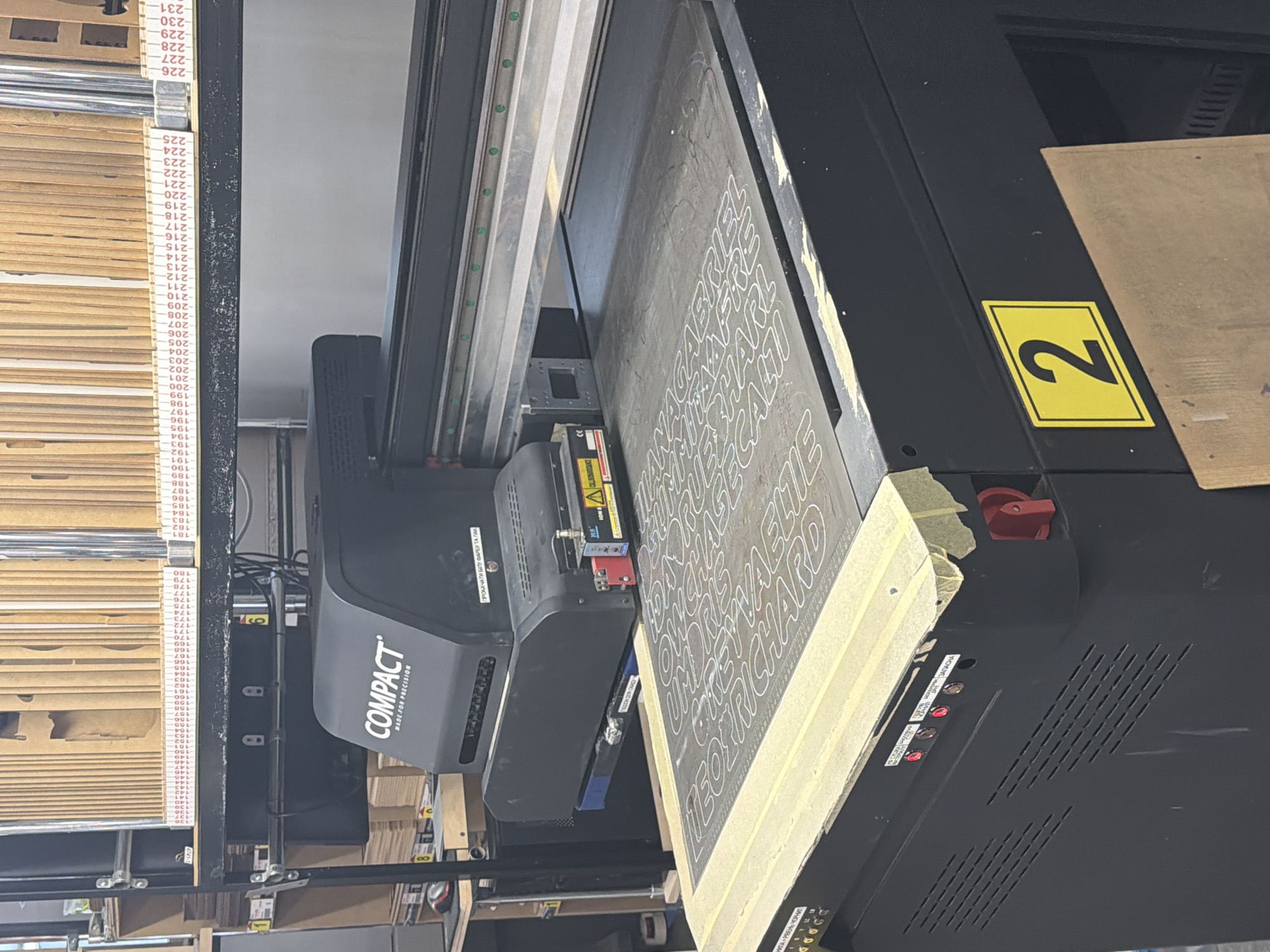 The image shows a flatbed printer in what appears to be a workshop or print shop. The printer is printing a design with white lettering on a dark surface. The name "RICHARD" is visible in the design. The printer itself is labeled "COMPACT MADE FOR PRECISION."
A yellow sign with the number "2" in black is affixed to the printer. To the left of the printer is a shelf unit with numbered compartments or slots, with numbers from "178" to "231" visible.
The label "ПРОКАЧАТИ БІЛУ ФАРБУ ТА ПАК" is visible. This text is in Ukrainian. The label "НИГА - РІВЕНЬ ЧОРНИГ" is visible, along with some Ukrainian labels describing the printer's operations.
The image shows a typical printing environment with equipment and organizational systems present. The precise location in Ternopil, Ukraine, cannot be determined from the image alone.