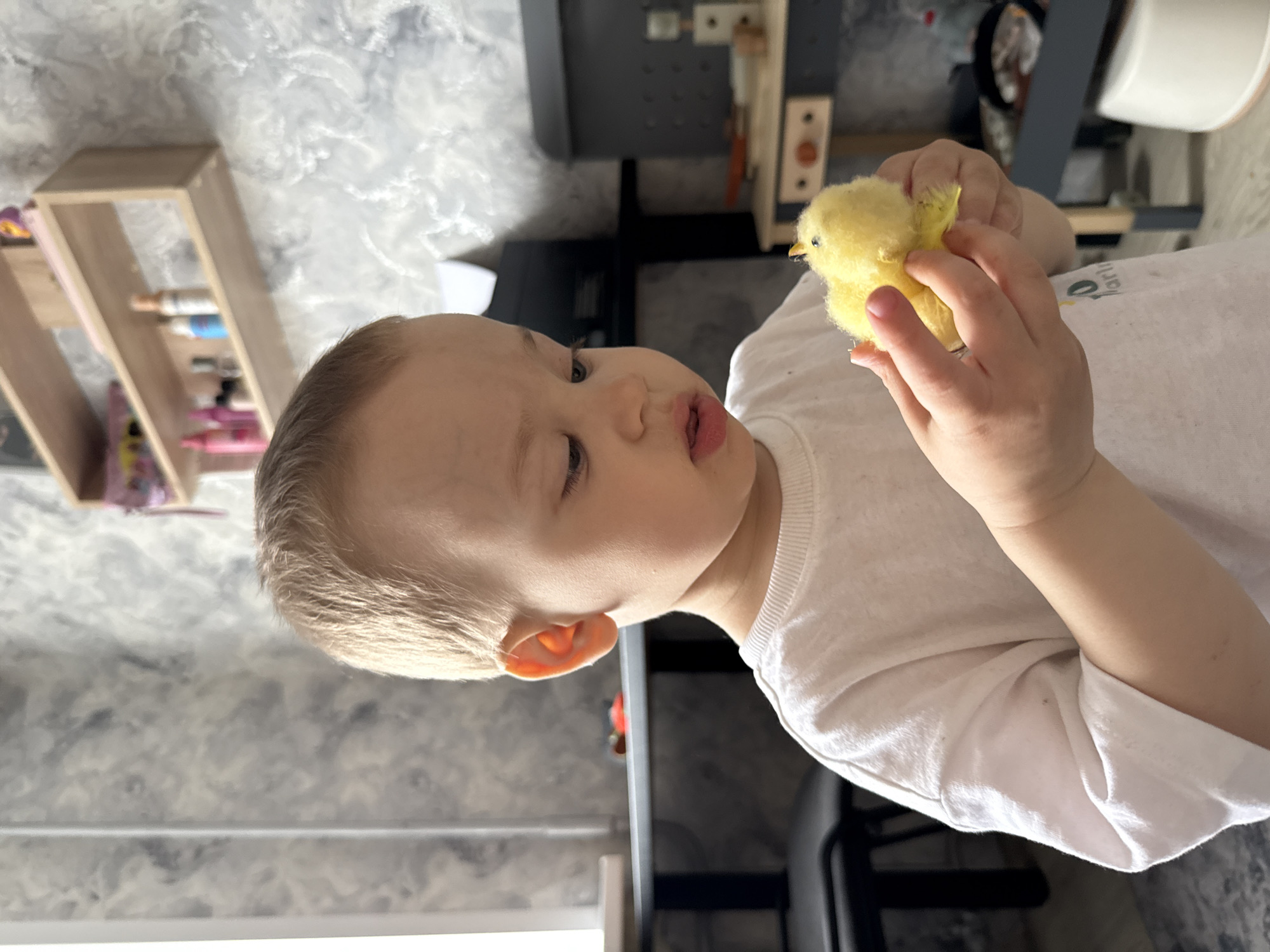 A young child with blond hair, wearing a white t-shirt, is holding a yellow chick in their hands. The background includes a grey wall, a wooden shelf with art supplies, and a black and wooden structure. The location is Unknown, Unknown.