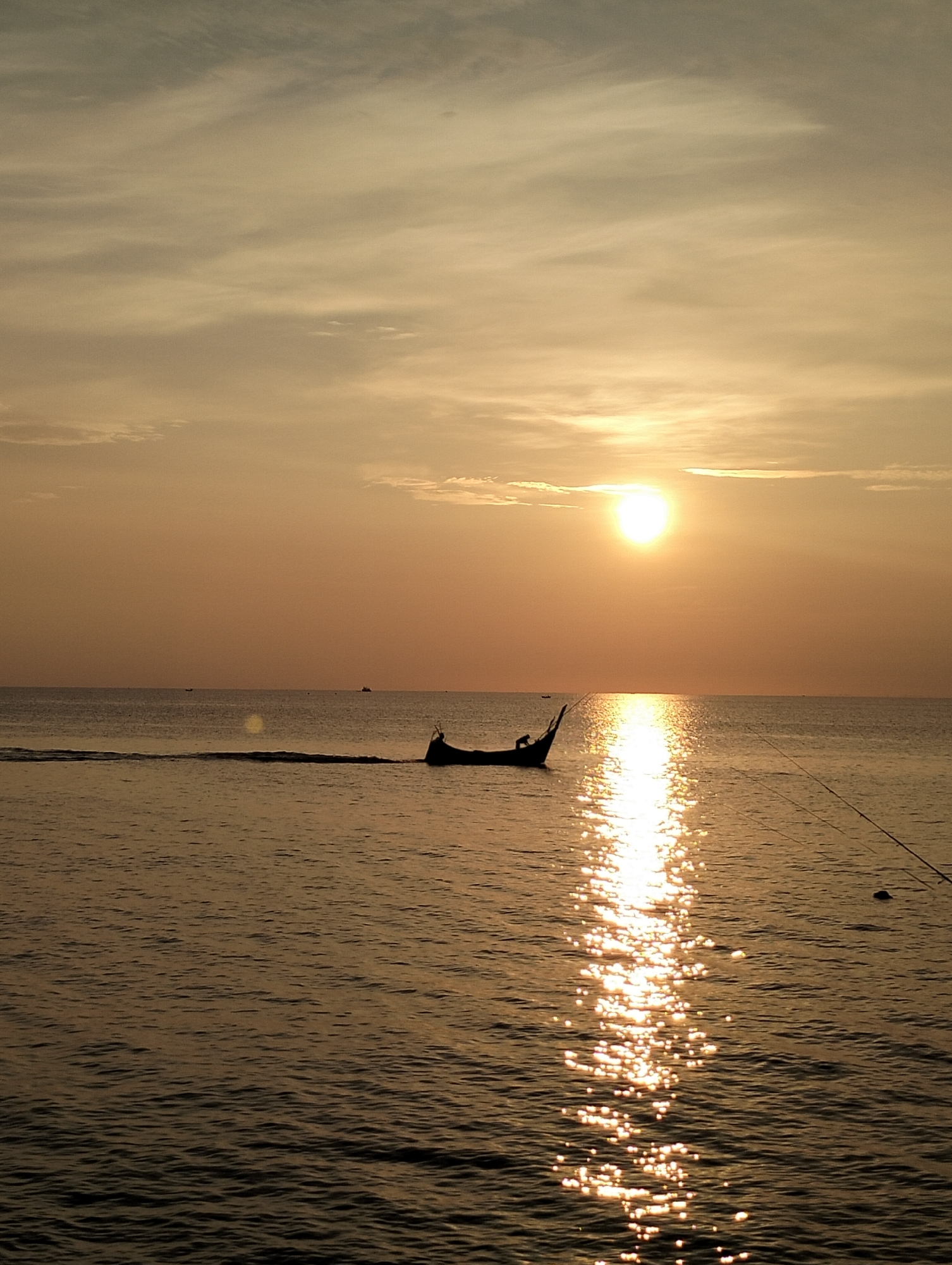 The image captures a sunset scene in Lhokseumawe, Indonesia. The sun is positioned in the upper-center, with its light reflecting off the water's surface as a bright, shimmering path. A small boat is silhouetted against the bright light of the sun, with a thin trail of water behind it. In the foreground, two fishing lines are visible. The sky is covered with clouds with an orange hue. The sea is calm, showing gentle waves.