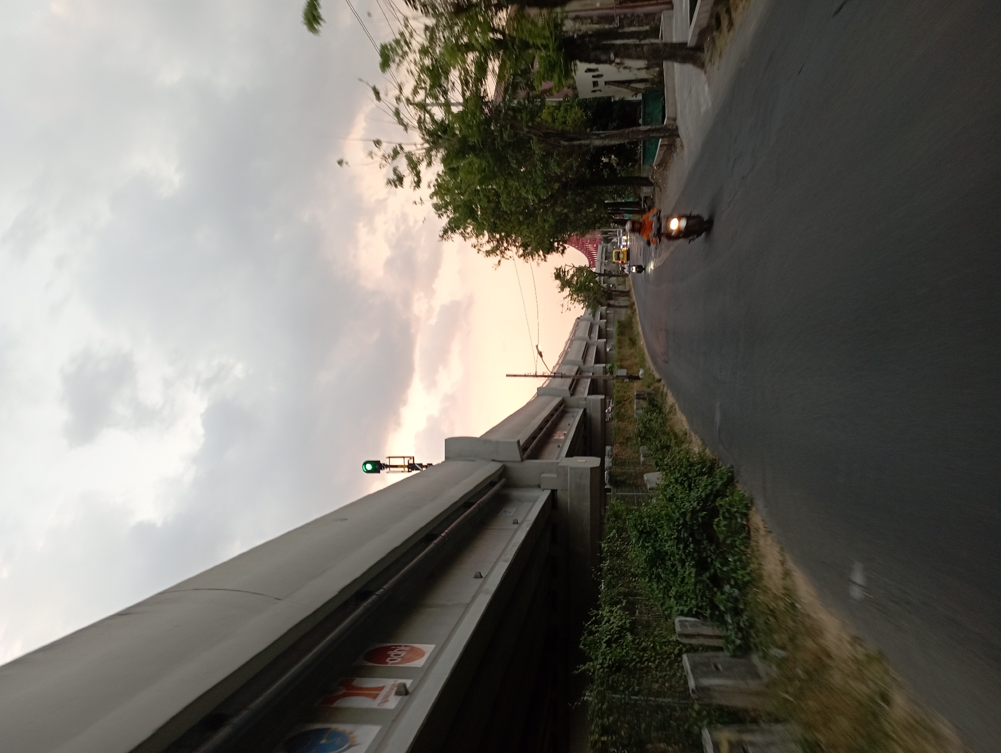 The image depicts an overpass structure in Surakarta, Indonesia, under a cloudy sky with the sun setting, casting a warm glow. The overpass, constructed of concrete, runs along the left side of the frame, with its underside visible. Several circular signs are attached to the side of the overpass. A green traffic light is illuminated on a pole extending from the overpass.
A paved road curves into the distance, with a motorcycle carrying a person in an orange shirt visible in the foreground, moving away from the camera. Further down the road, another motorcycle and a yellow vehicle are also in motion. Lush green foliage grows along the base of the overpass and on the right side of the road, interspersed with trees and the facades of buildings. The scene appears to be captured at dusk, as indicated by the soft light and the illumination from the motorcycle's headlight.