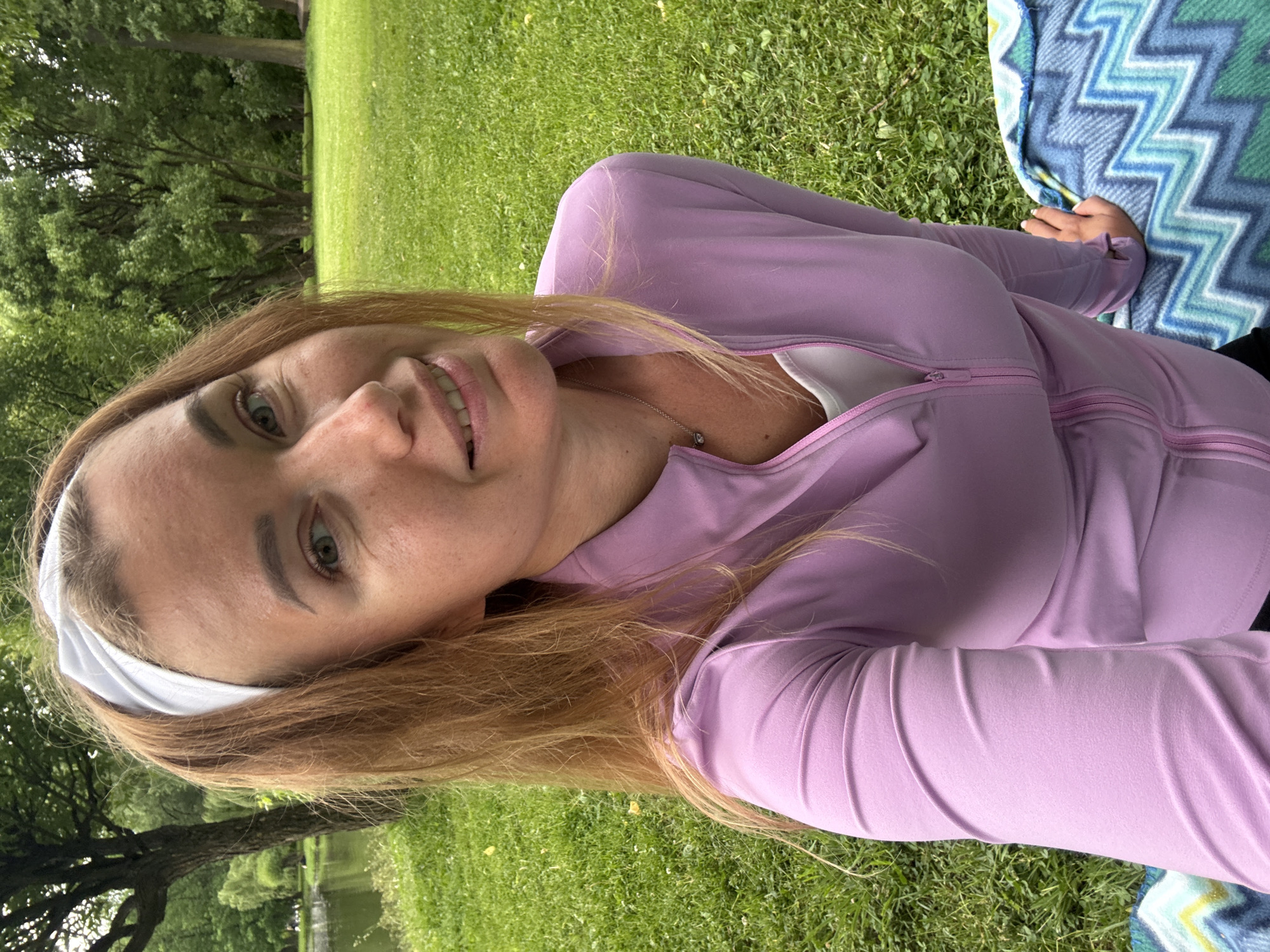 A medium shot shows a woman with long, reddish-blonde hair, wearing a white headband, a pale pink zip-up jacket over a white top, and a silver necklace with a small pendant. She is smiling and looking at the camera. She is seated on a blanket with a blue, green, and white zig-zag pattern. The background is a grassy park with trees and a body of water on the left side. The lighting is natural and the image is taken outdoors.