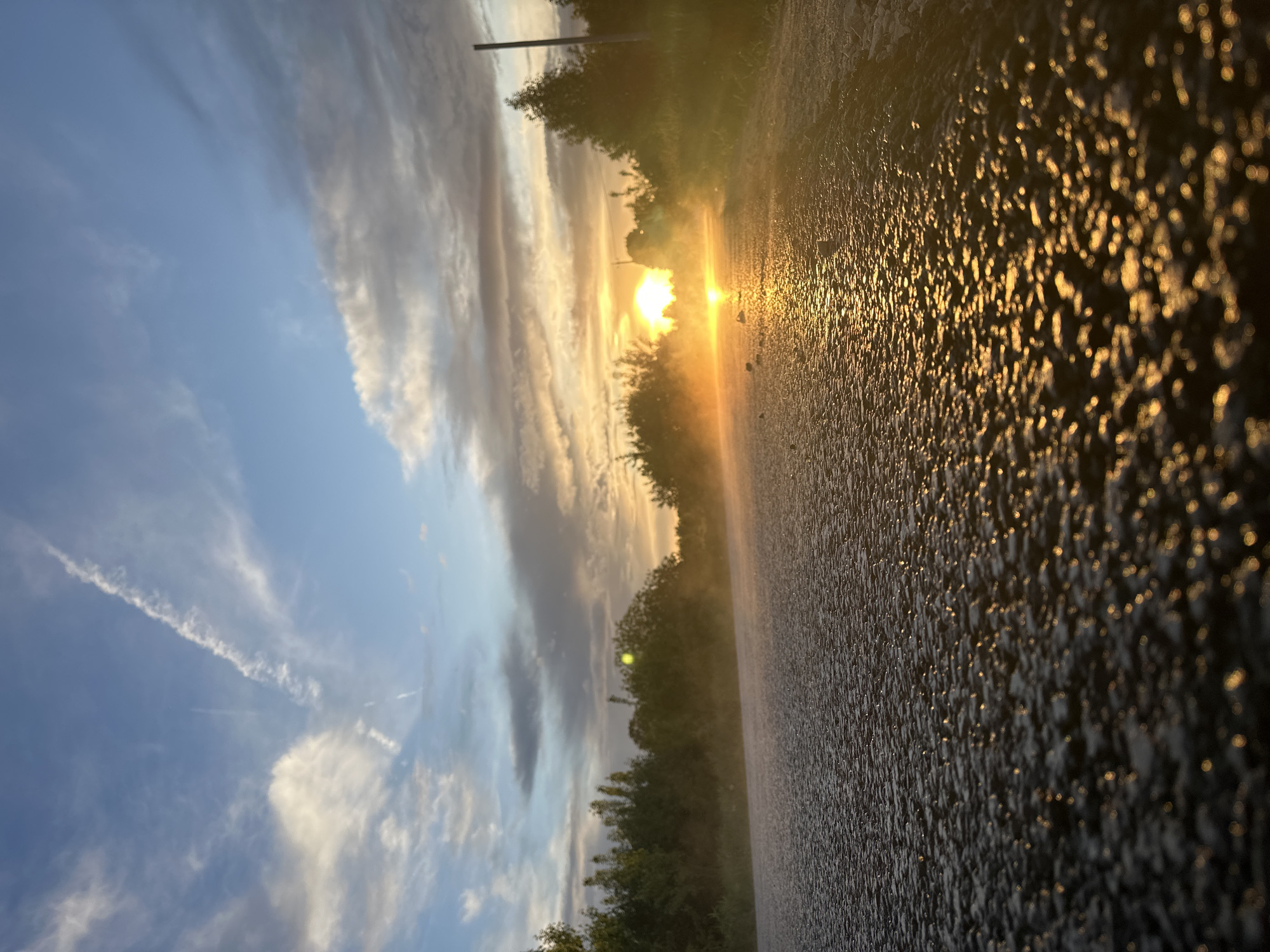 The photograph captures a low-angle perspective of a road leading towards a setting sun. The road surface, characterized by a rough texture, occupies a significant portion of the foreground, with its reflective sheen suggesting it may be wet or recently washed. The sunlight glints off the road, creating a mosaic of bright spots.
Beyond the road, the setting sun is positioned between a line of trees, its intense light partially obscured, producing a radiant glow. The sky above is a mix of blue and soft hues, with wispy clouds stretched across. The overall composition is tilted to the side.
The location appears to be in a place with trees lining a road, and without additional information, definitively pinpointing it to Shchyolkovo, Russia, is not possible.