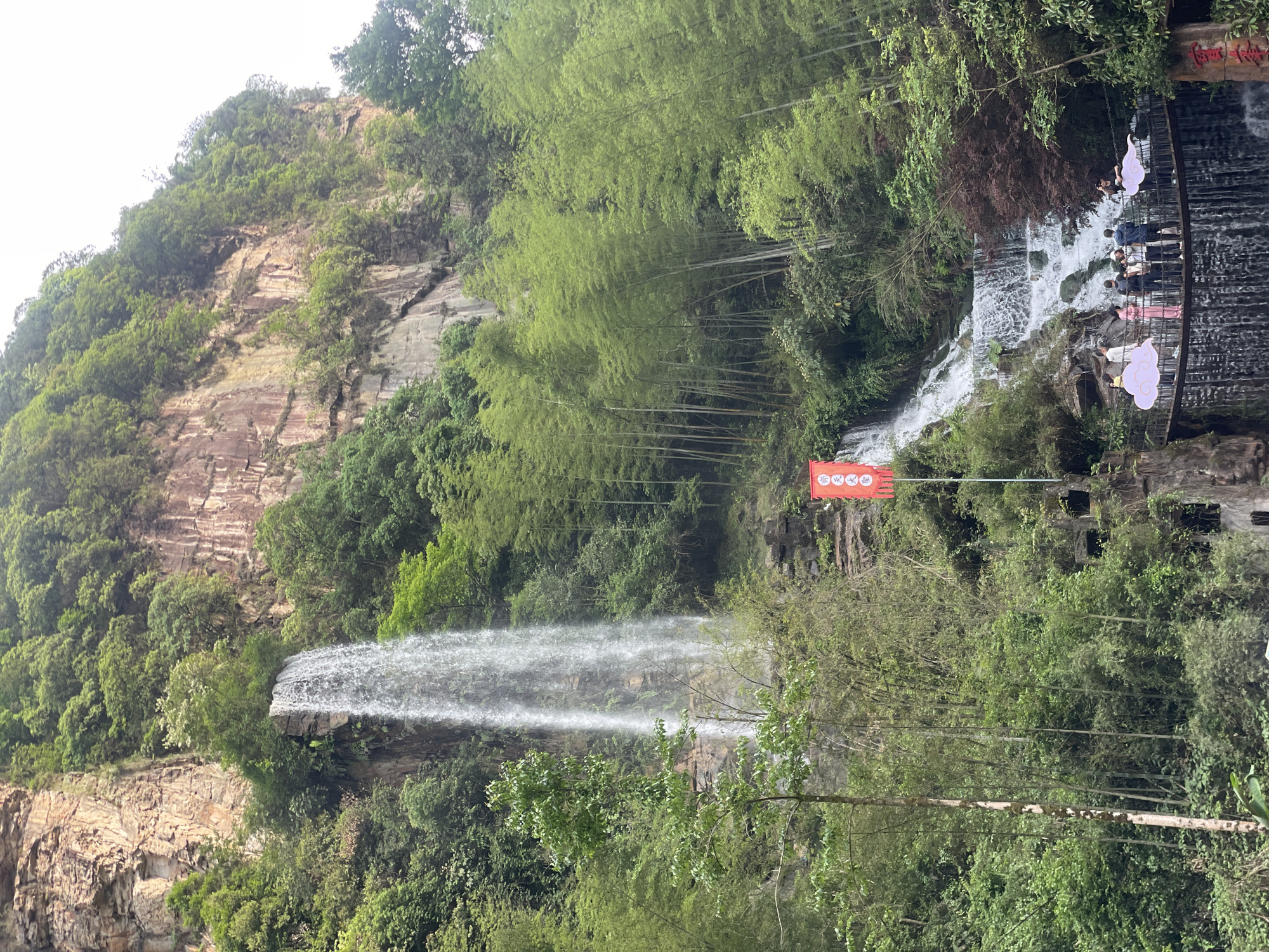 A landscape view features two waterfalls framed by lush vegetation in Wulingyuan, China. The primary waterfall cascades from a high, rocky cliff face covered in greenery on the left side of the frame. To the right, a smaller waterfall flows under a decorative walkway where people are standing. A red flag with Chinese characters hangs near the walkway. Bamboo trees are prominent in the background. The weather appears overcast.