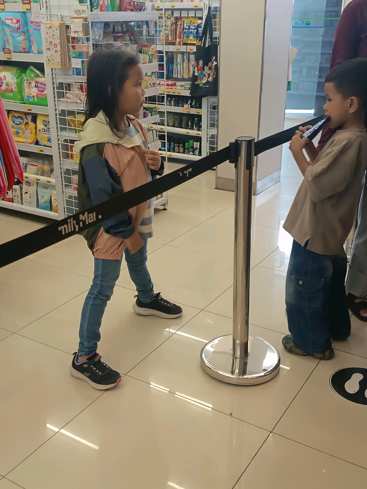 This is a medium shot of two young children standing in a retail store, likely in South Jakarta, Indonesia. A young girl with dark hair is standing on the left, facing right, wearing a light-colored hooded jacket over a striped shirt and blue jeans. A younger boy with short dark hair is on the right, facing left and looking down at a black object he is holding to his mouth. He is wearing a light brown t-shirt and baggy denim jeans. Both children are standing behind a retractable belt barrier. The environment appears to be a store aisle with shelves stocked with various products visible in the background. The floor is tiled and reflects the overhead lighting.