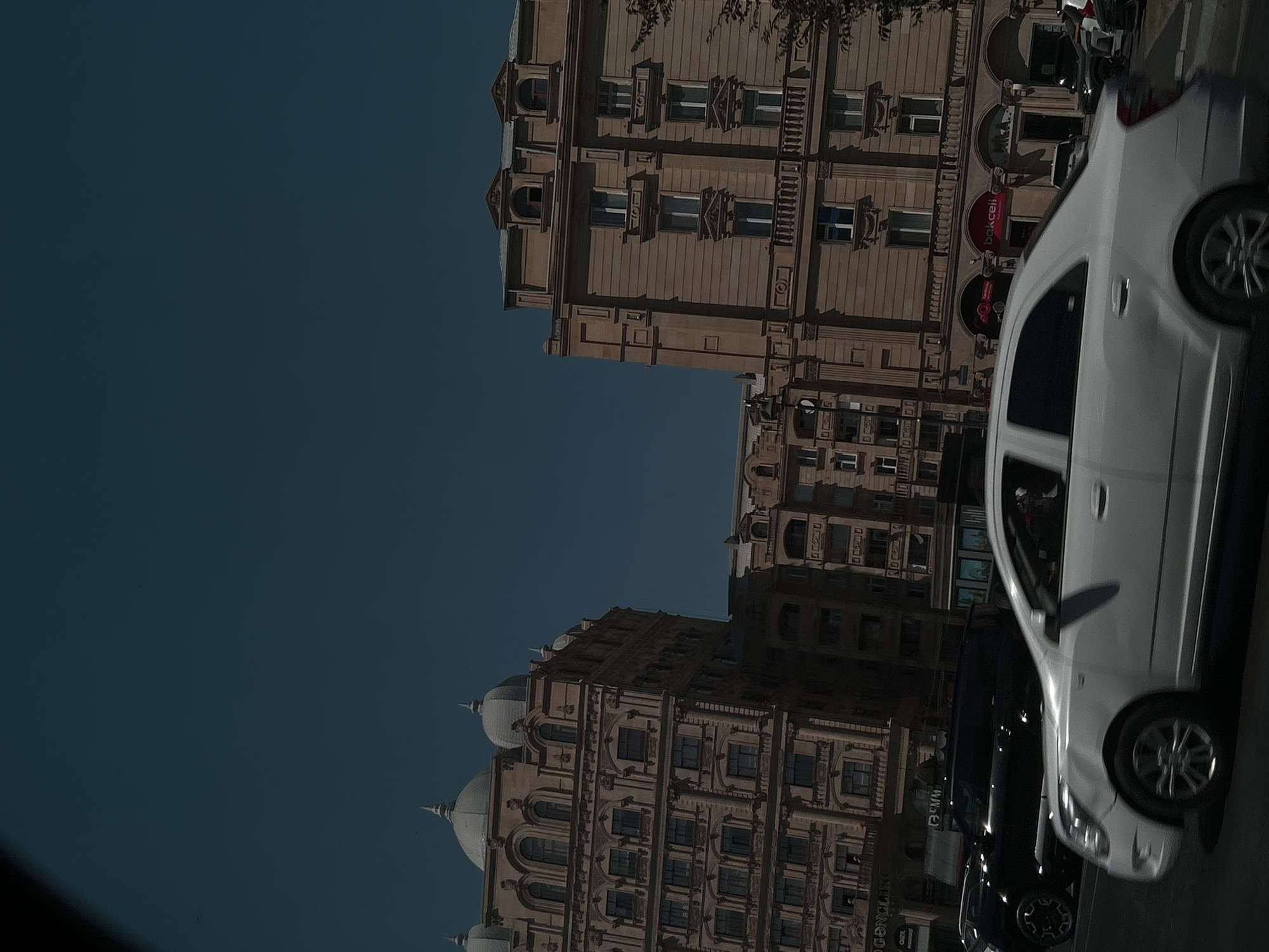 The image shows a street scene in Baku, Azerbaijan, with a bright blue sky overhead. In the foreground, a white sedan is driving on the right side of the road, partially obscuring a black SUV behind it. The buildings lining the street are ornate and feature classical architecture, with arched windows and decorative facades. The building on the left has multiple domes, and the building on the right has a multi-story facade with balconies. Businesses are visible on the ground floor of the buildings, with signs in Cyrillic and Latin script. The overall impression is of a well-established urban environment.