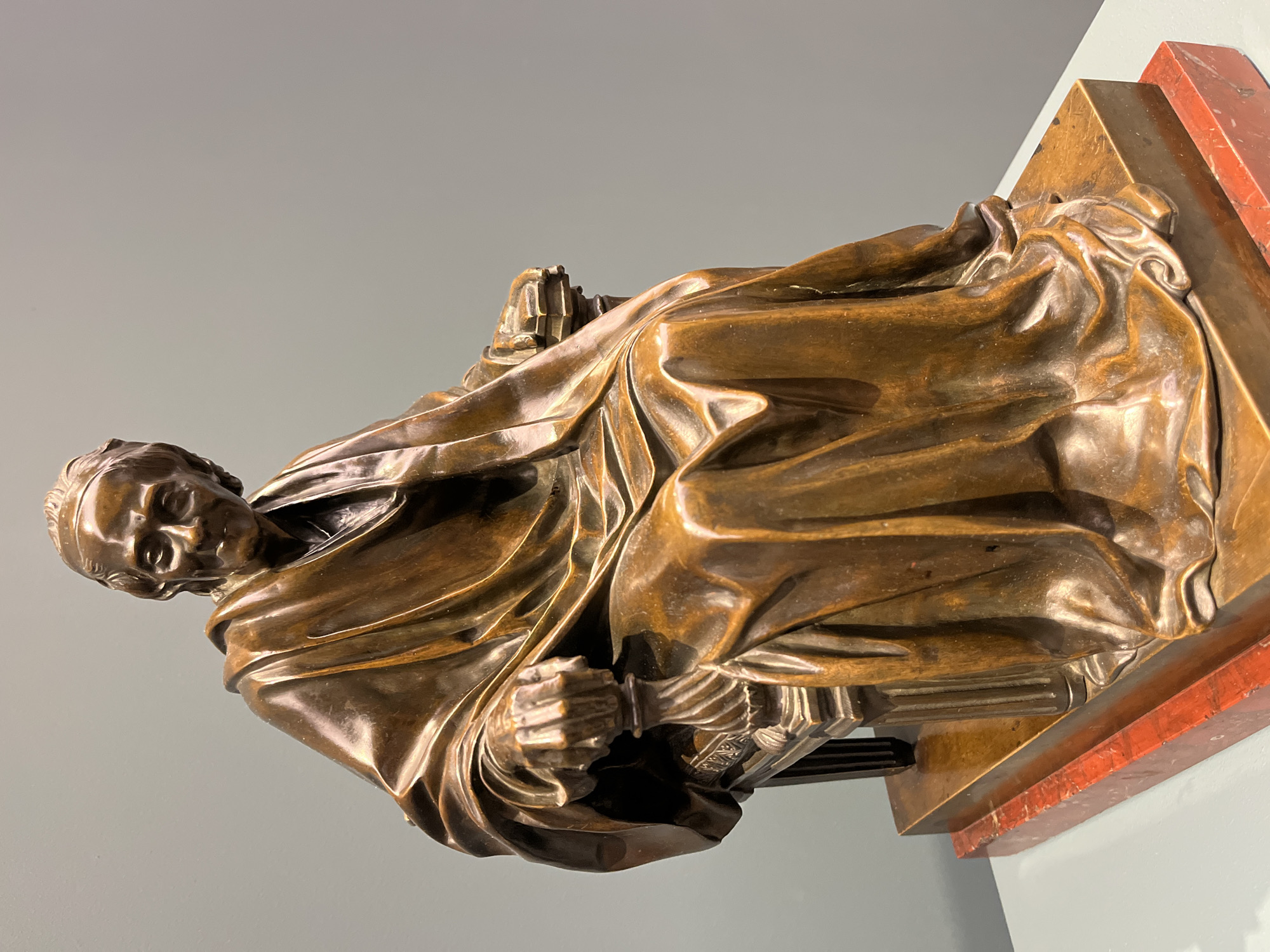 A bronze statue of a person in a cloak is positioned on a brown and red base against a grey wall. The location is Unknown, Unknown. The person depicted is holding what appears to be a book or a similar object.