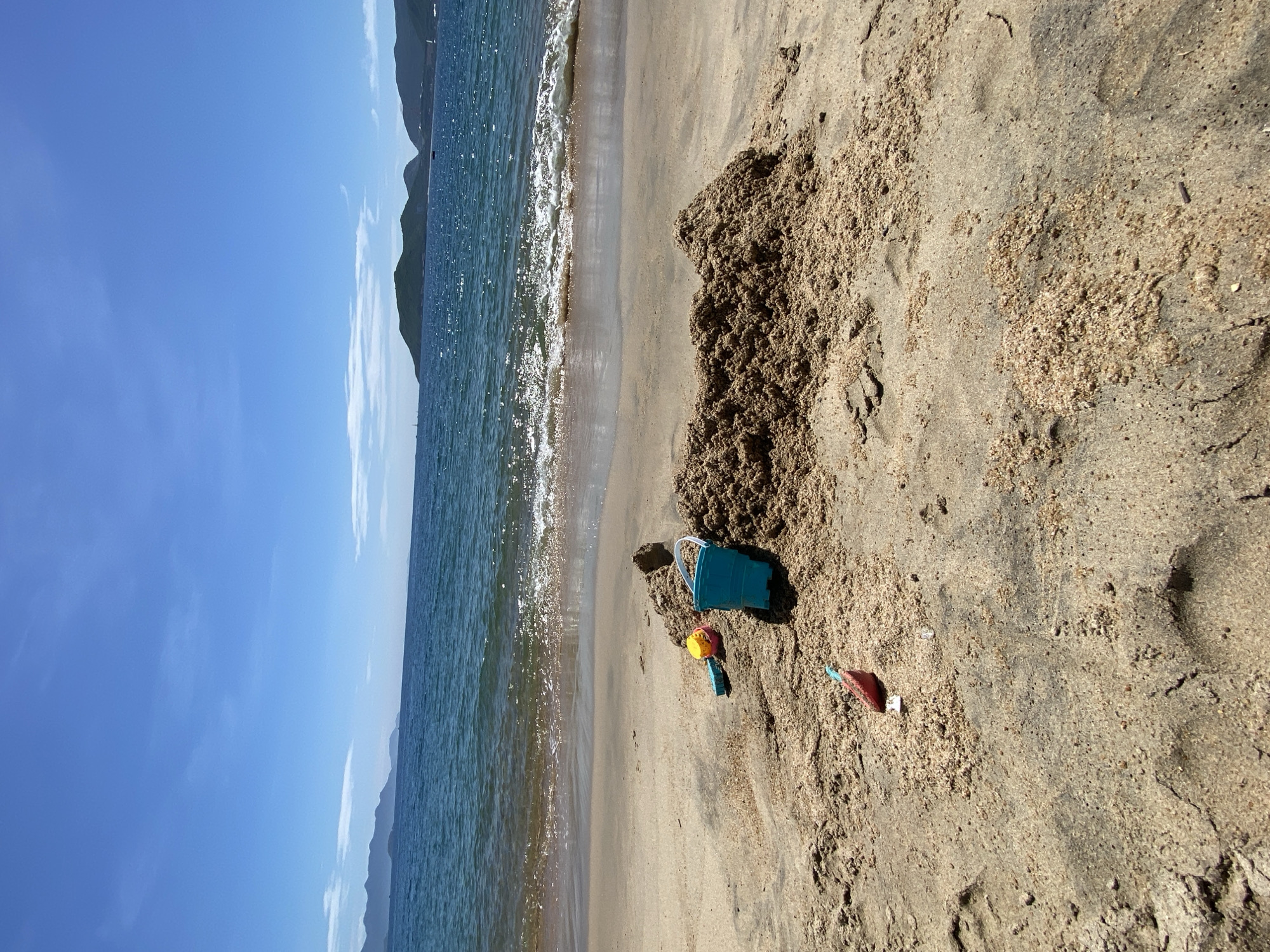 The image captures a tranquil scene on a sandy beach in Nha Trang, Vietnam. The foreground is dominated by a pile of damp sand, suggesting recent activity in building a sandcastle. Scattered around the sandcastle are children's beach toys: a small blue bucket, a yellow shovel, and a red spade. The toys are positioned on the sand, some partially buried, indicating they have been used.
Beyond the beach, the tranquil turquoise waters of the sea extend towards the horizon. Gentle waves with white foam are washing ashore, creating a subtle line of wet sand where the water meets the beach. In the distance, the silhouette of a chain of verdant, mountainous islands rises from the sea under a clear, bright blue sky with scattered wisps of white clouds. The overall atmosphere is one of a peaceful, sunny day at the beach.