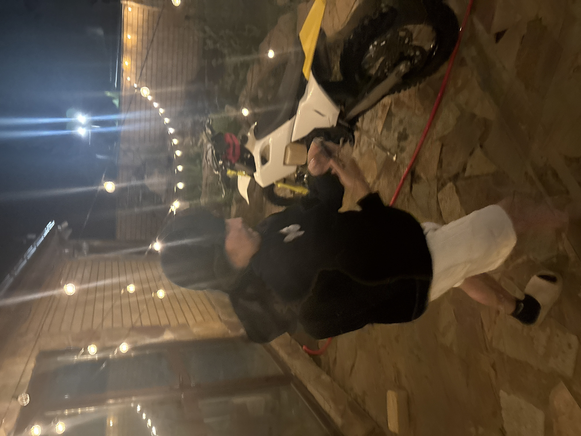 This late evening or nighttime image, taken in Orekhovka, Russia, captures a casual outdoor scene illuminated by string lights. The photograph is oriented vertically, showing a person standing next to an off-road motorcycle on stone-paved ground.
The main subject is a male figure, standing towards the left side of the frame (when corrected for rotation). He is wearing a dark, possibly black, hoodie with a white emblem on the chest, white shorts, and black and white slides. He appears to be holding a beverage or small object in his right hand. A dark helmet or hat is visible on his head. His gaze is directed towards the left.
To his right stands a white and yellow dirt bike or off-road motorcycle, prominent in the frame. Its dark tires and some mechanical components are visible. A red rope or cable lies on the ground near the person's feet and extends across the scene.
The background features a building or wall constructed from light-colored bricks or wooden planks, brightly illuminated by numerous round string lights hanging in rows. These lights cast a warm glow across the scene, creating a festive ambiance. The ground is made of irregular stone pavers, suggesting a patio or garden area. In the bottom left, a reflective surface, possibly glass, shows distorted reflections of the string lights. The overall atmosphere is relaxed and informal. No discernible text is visible on any objects or surfaces.
