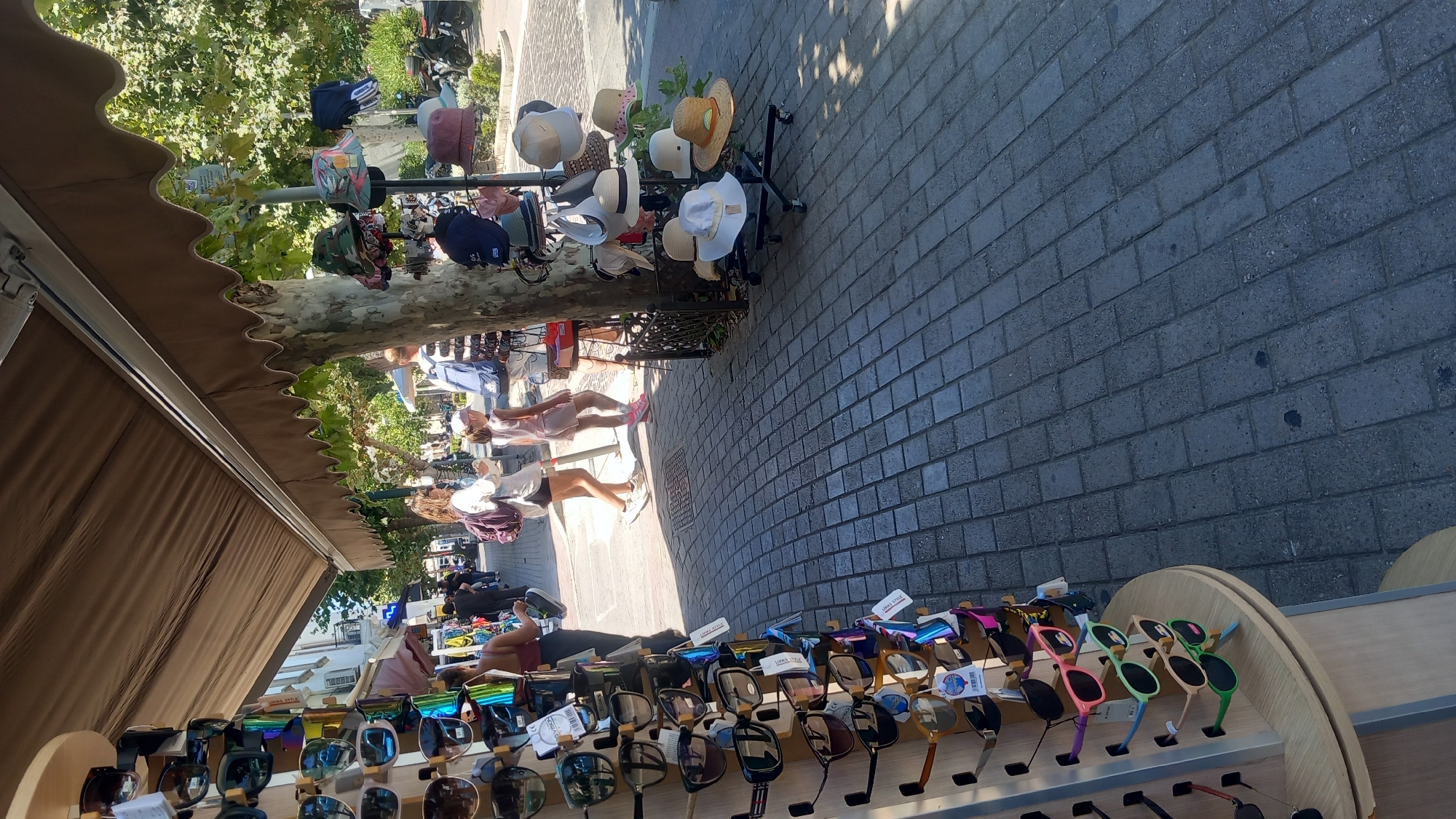 The image shows a sidewalk scene in Kos, Greece, likely in a tourist area given the merchandise displayed. In the foreground, a display stand showcases a variety of sunglasses on the left, with different colors and styles. To the right, a rack holds several hats, including straw hats and bucket hats, some of which are being sold from a rolling stand. A tree with green foliage provides shade over a portion of the scene. In the background, several people are walking along the sidewalk, including two women, one carrying a backpack, and a child wearing a pink hat and dress. The sidewalk is paved with grey interlocking bricks. The overall atmosphere suggests a sunny day in a bustling commercial area, possibly near the beach or a promenade.