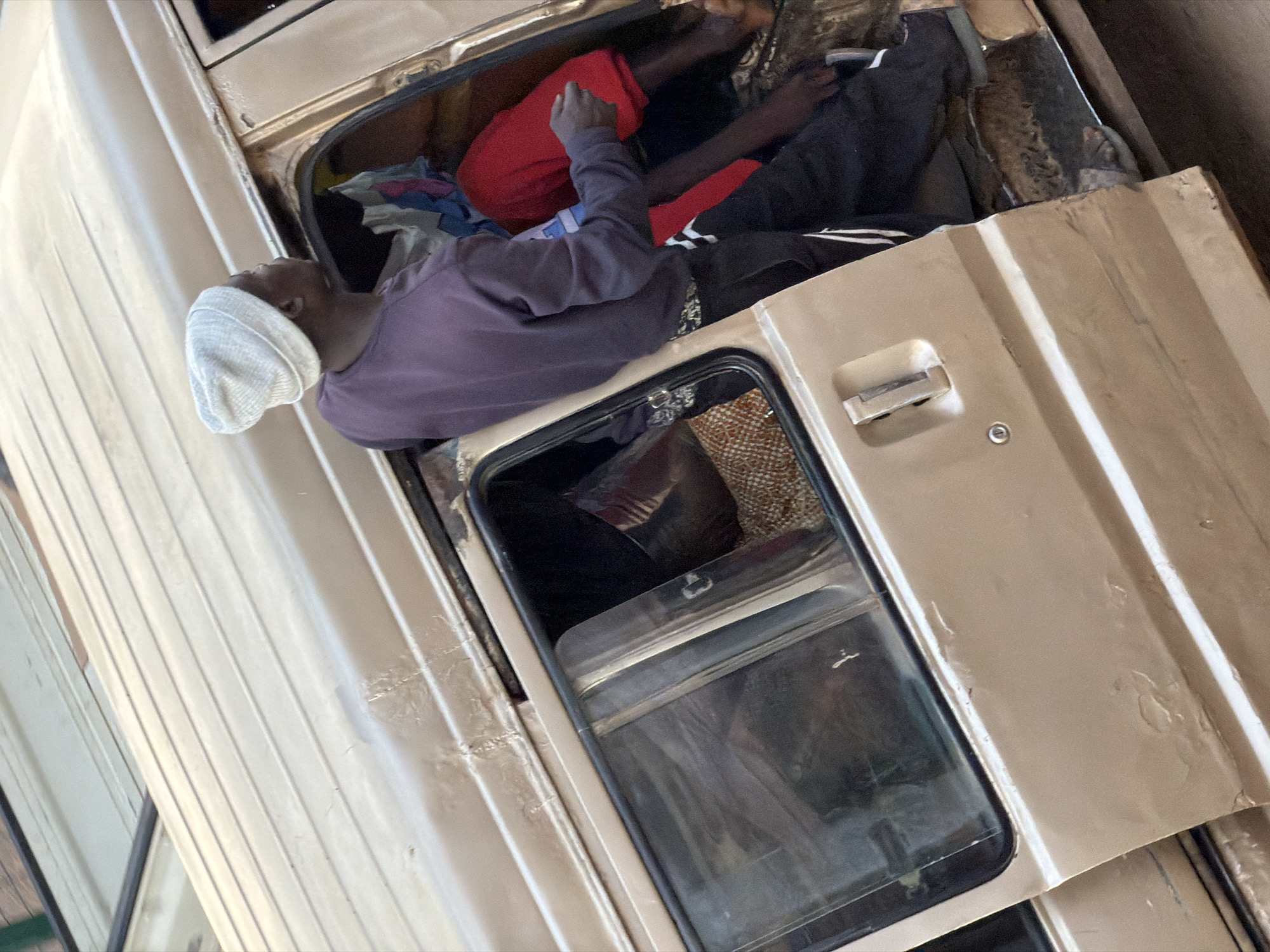The image shows the interior of a slightly damaged, tan-colored van in Bukuru, Nigeria. A person wearing a purple long-sleeved shirt and black pants with white stripes, and a white beanie hat, is standing in the open doorway of the van, looking to the right. Inside the van, another person is visible, wearing a red garment. Several other items, including what appears to be fabric and a patterned textile, are also inside the vehicle. The van's exterior shows signs of wear, including dents and peeling paint.