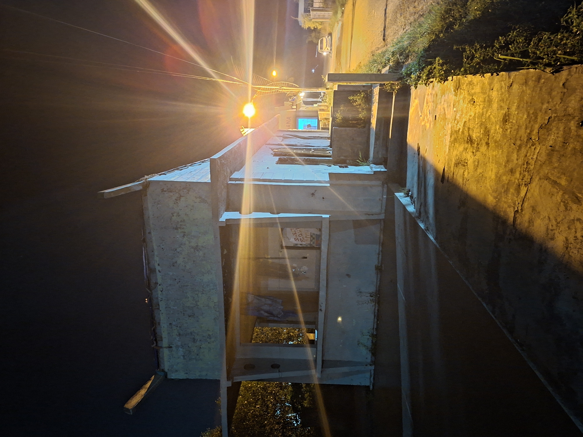 This photo, taken in Lhokseumawe, Indonesia, captures a nighttime scene with a strong, centrally positioned light source causing lens flare. The main subject is a weathered, concrete structure with multiple openings, resembling an abandoned building or bus stop. The structure's interior reveals glimpses of foliage and possible stored items. To the right of the concrete structure, a textured concrete wall is illuminated by the light source. The wall has a rough surface and appears to be aged. Vegetation grows along the top of the wall, indicating the location is outdoors. In the background, across the street, there's a glimpse of buildings and a vehicle. The left side of the image is primarily dark, suggesting the time of day or location away from the light source. The image's composition, lighting, and subject matter suggest an urban or peri-urban environment in disrepair.