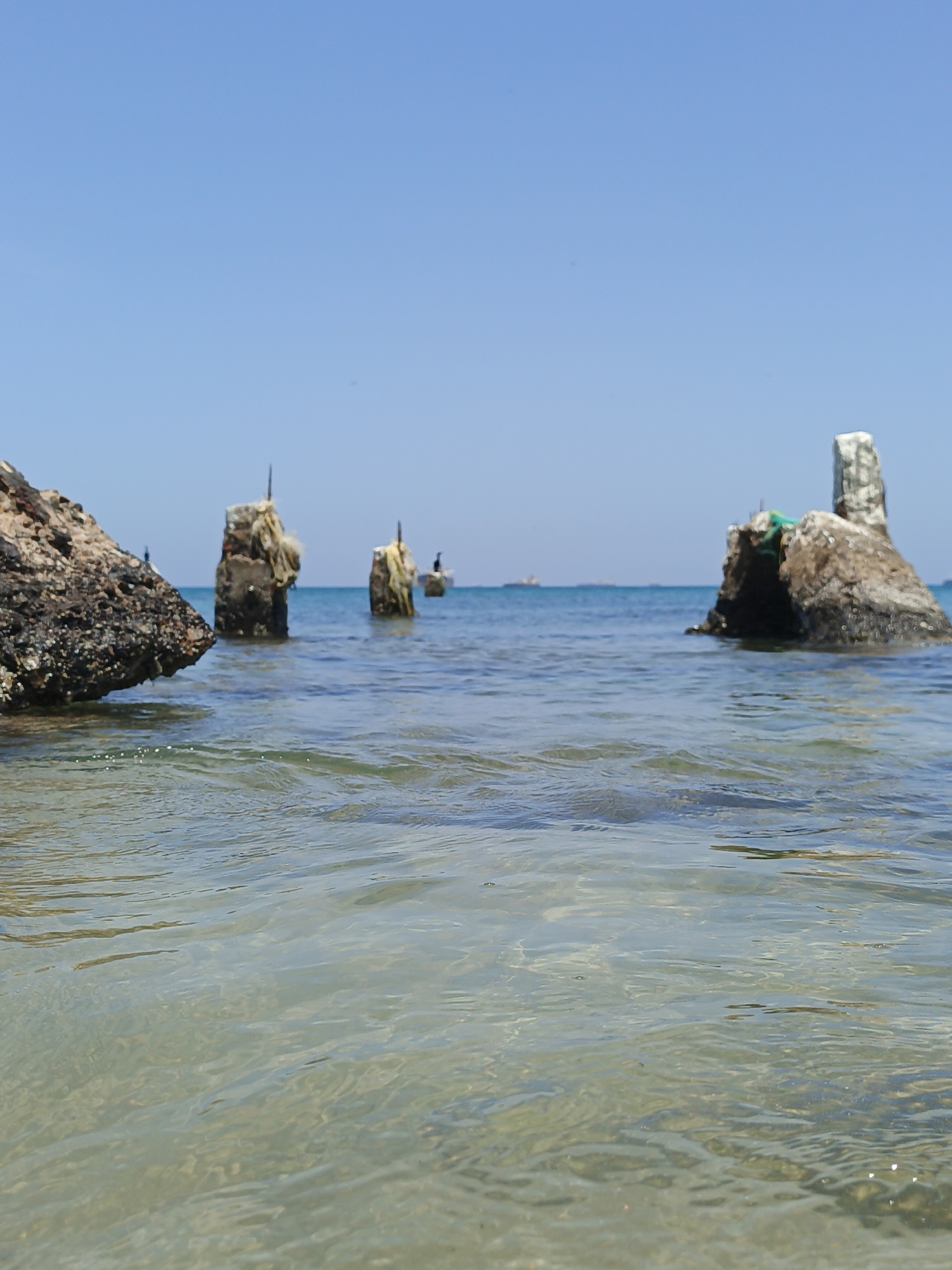 The scene depicts a shoreline in Villa Marina, Venezuela. The foreground features clear, shallow water revealing a sandy bottom. Partially submerged concrete and rock structures are scattered throughout the water. Some of these structures appear to be remnants of old pylons or breakwaters. A few birds perch on some of these structures. The horizon shows a distant skyline with what appears to be ships or other vessels. The sky is clear and blue, suggesting a sunny day. There are no people visible in the scene.
