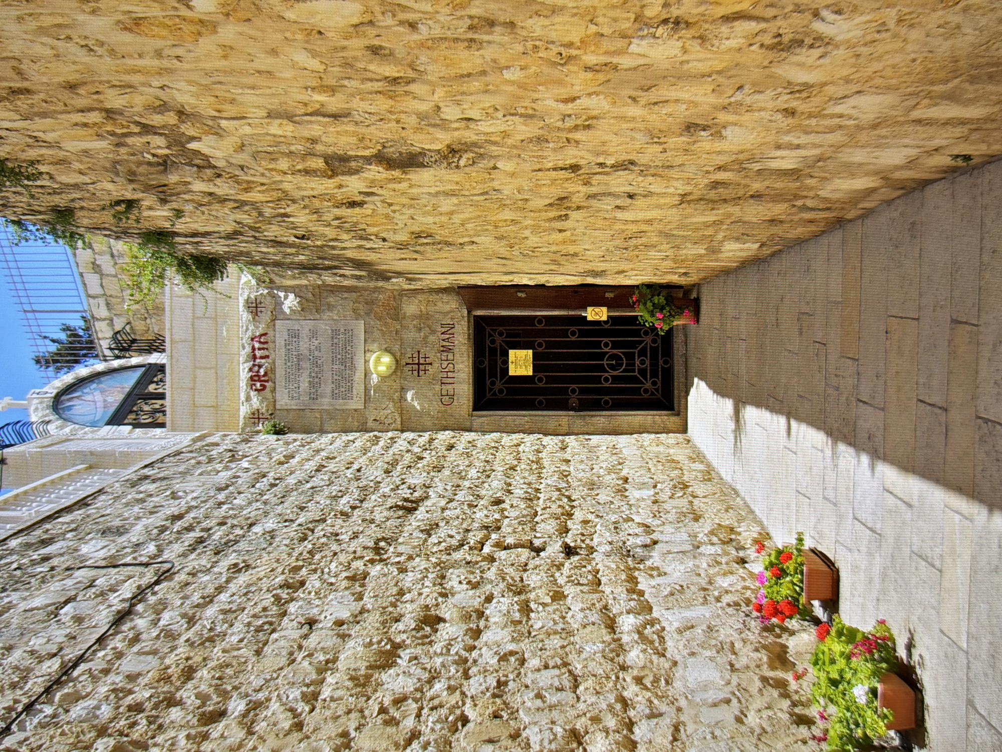 The image depicts a narrow stone alleyway with tall, rough-hewn stone walls on either side. The alley leads to a dark metal gate with ornate scrollwork, set into the right-hand wall. Above the gate, there is a sign with text and a stylized cross. The ground of the alley is paved with large, light-colored stone tiles, and a strong shadow is cast across it from the left.
On the left side of the alley, near the foreground, are two terracotta pots filled with flowering plants. The plants have green leaves and clusters of red and pink flowers.
On the right side of the alley, closer to the gate, is another terracotta pot with flowering plants, also with green leaves and pink flowers.
The walls are constructed of irregular, light brown and tan stones. The wall on the left appears to have some darker lines or marks, possibly from wires or conduits.
In the background, at the end of the alley, the alley opens slightly, and there is a glimpse of a building with white trim and an arched window with decorative metalwork. Above this, a blue sky with a metal fence is visible.
A sign with red lettering is visible on the stone wall on the right, above the gate, which reads "CROZIA" and below it, some text in smaller print. Further down on the right wall, there is a yellow sign attached to the gate.
The overall impression is one of a historic, possibly religious, site. The stone construction, religious symbols, and the name "Gethsemani" inscribed above the gate suggest a connection to biblical places.
The location is identified as the city of Kyiv, Ukraine, though this specific scene does not contain any obvious Ukrainian architectural or cultural identifiers.